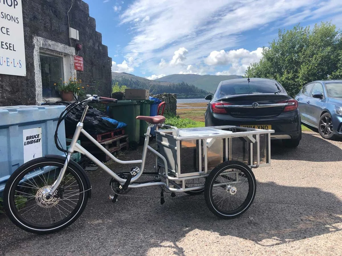 A cargo bike with a metal frame and two wheels parked on a gravel lot, with two cars and a stone building in the background, overlooking a lake and mountains under a partly cloudy sky.