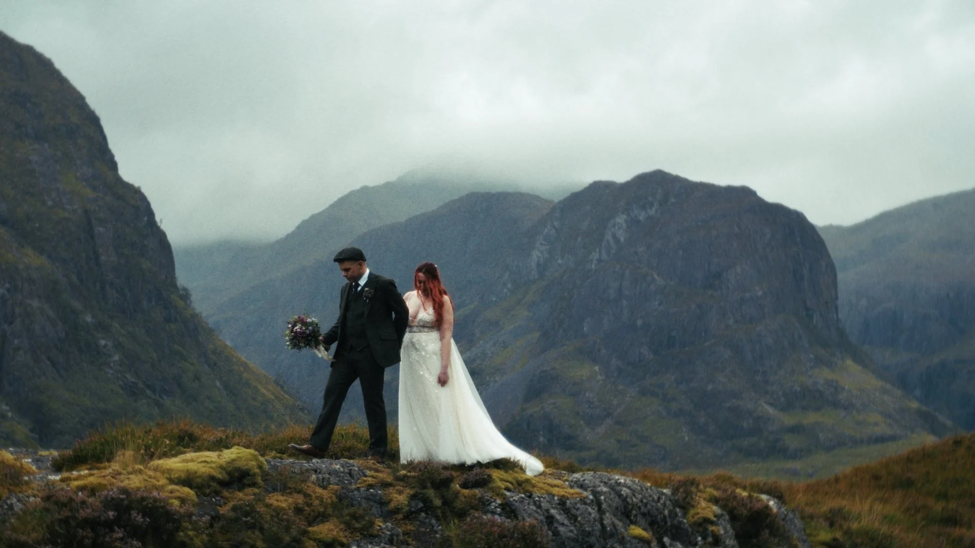 A bride and groom walking together in a mountainous landscape, with the bride holding her dress and the groom holding a bouquet of flowers, under an overcast sky.
