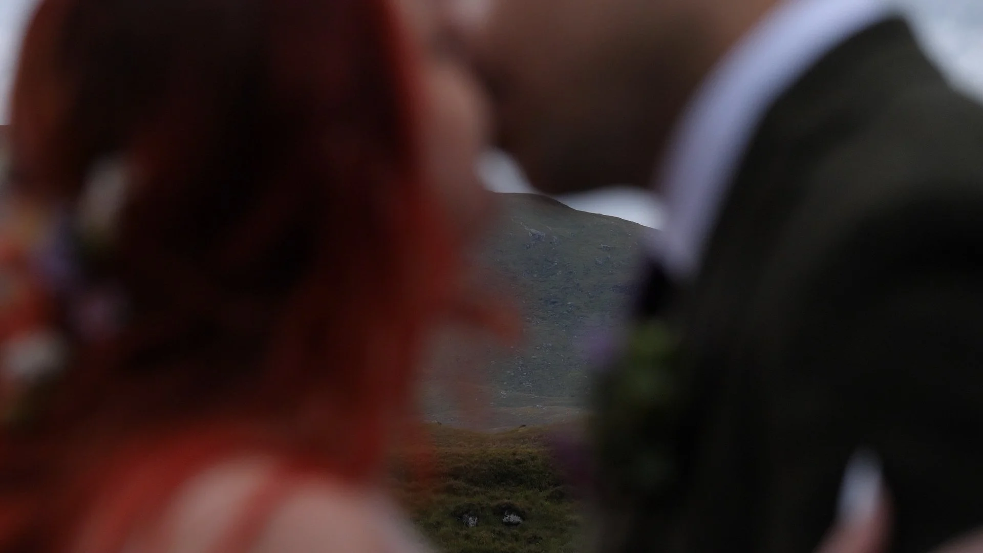 A blurry person with reddish hair and a suit, with a mountain landscape in the background.