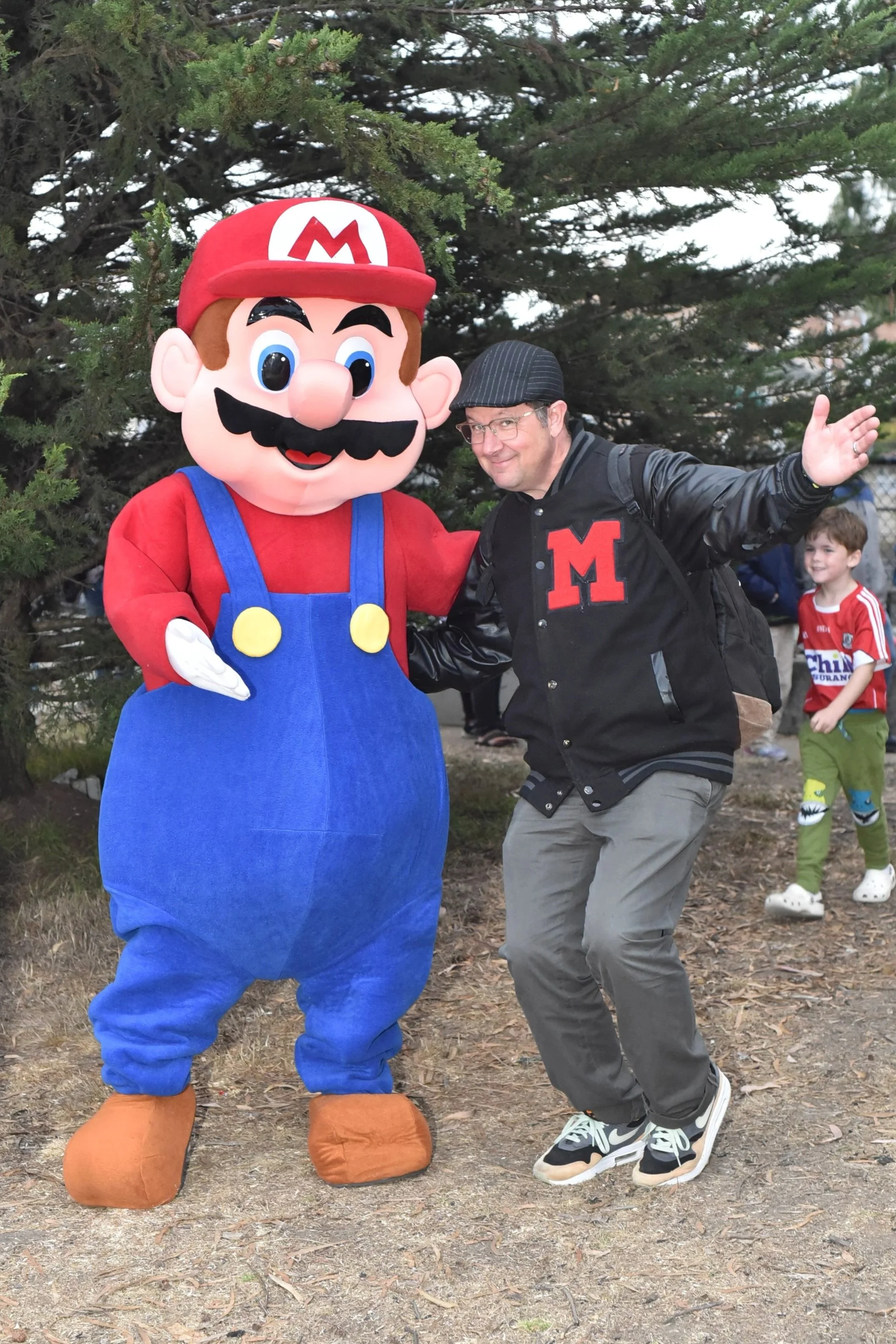 A man posing with a Super Mario mascot character from video games. They are outdoors near a tree, with other children visible in the background.