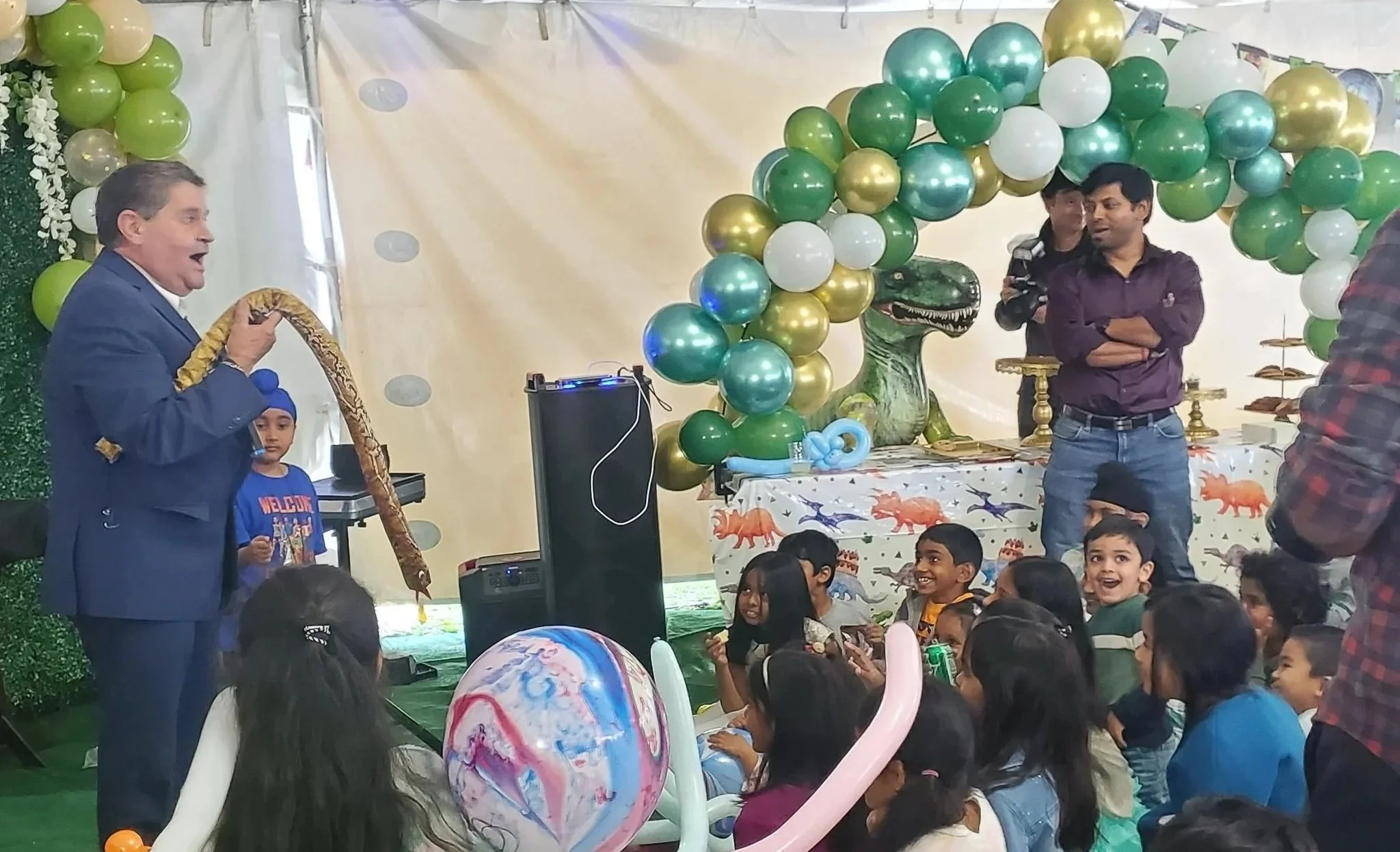 A birthday party scene with children and adults smiling at a magician, balloon animals and gold and green balloon decorations, a dinosaur-themed tablecloth, and a magician in a blue suit holding a snake. Children appear happy, seated and engaging.