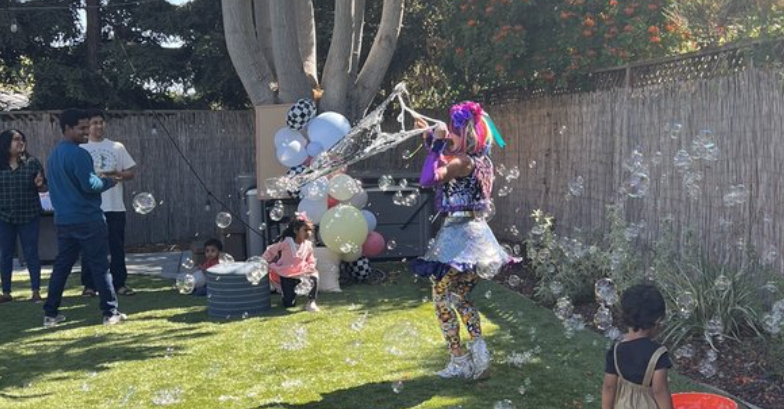 Entertainer in sparkly skirt and butterfly top makes giant bubbles at a child's birthday. Parents are smiling.