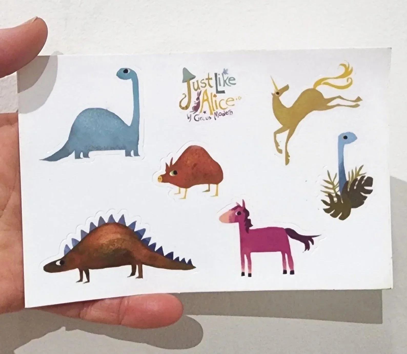 Colorful stickers of various dinosaurs and the words 'Just Like Alice' with smaller text 'by Circus Modern'.