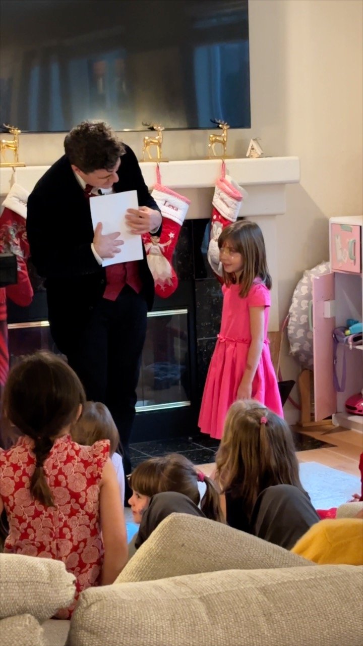 A magician dressed in a tuxedo presents a gift to a young girl in a pink dress during a holiday gathering in a living room decorated with Christmas stockings and ornaments. Several children are seated engaged in the magic show and the birthday girl s