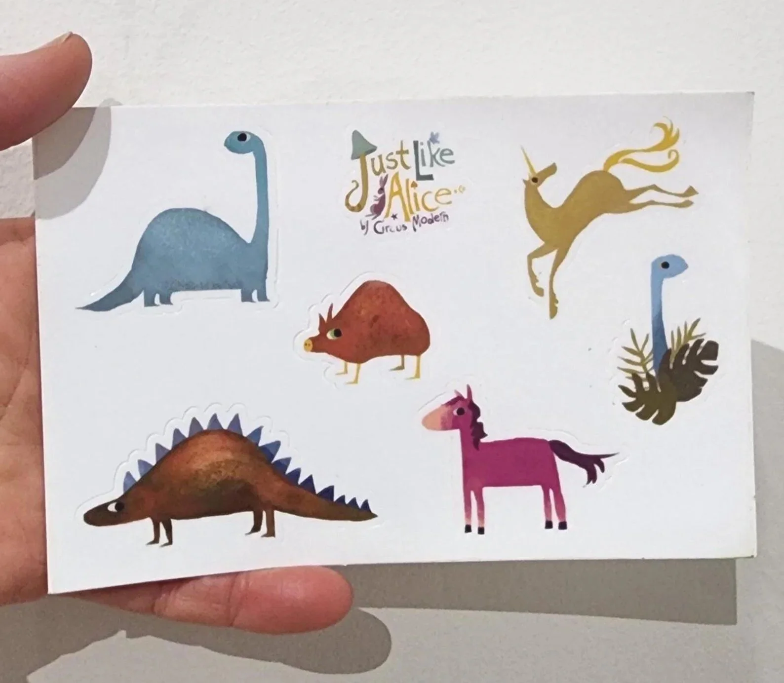 Colorful dinosaur, unicorn, and animal stickers on a white sheet  with the text 'Just like Alice Circus Modern' in the center.