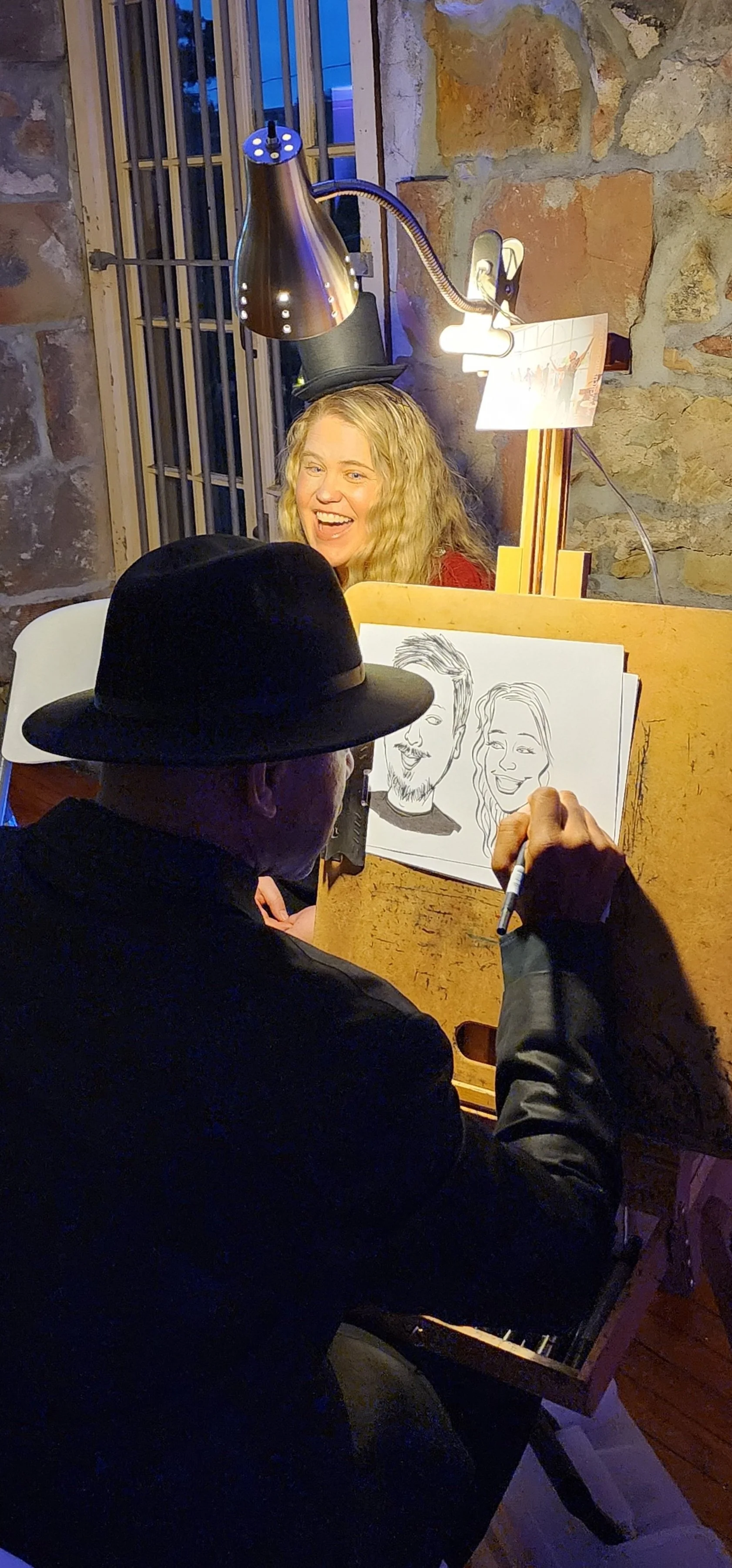 A man wearing a black hat and jacket drawing caricatures of a woman with curly blonde hair and a man with a beard, as she smiles in the background with a top hat on her head and electric lamp overhead.