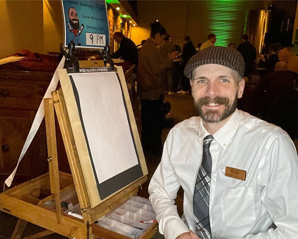 A smiling man wearing a white shirt, tie, and apron, sitting at a table in a restaurant next to an empty artist's easel and a sign indicating a drawing event at 9 PM. The background shows a busy, dimly lit restaurant interior with patrons.