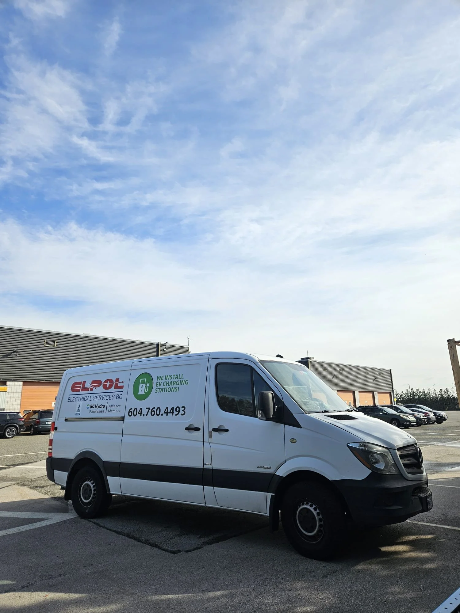 White service van parked in a parking lot with multiple cars, a building with a dark roof in the background, and a partly cloudy sky.