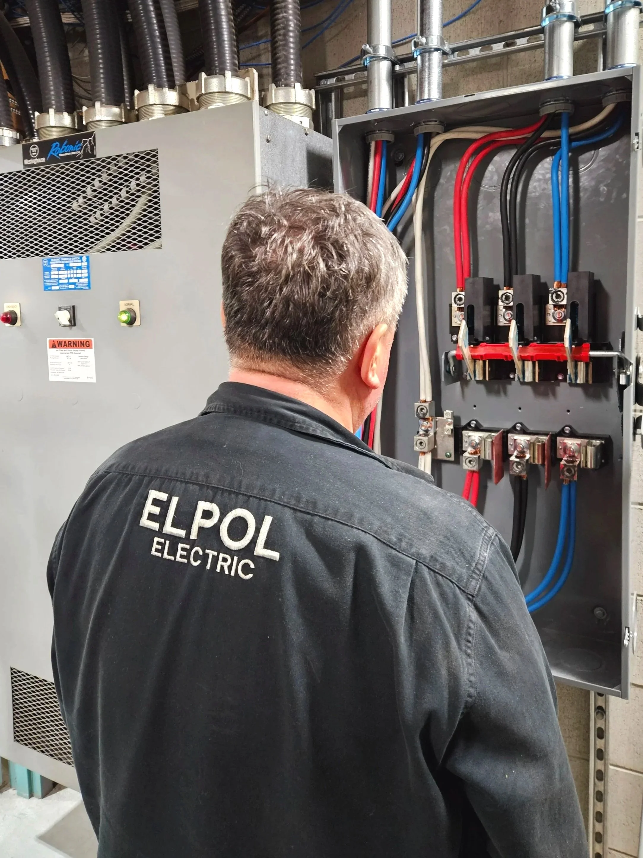 An electric technician working on an electrical panel with colored wires, switches, and circuit breakers, wearing a black jacket with 'ELPOL ELECTRIC' embroidered on the back.