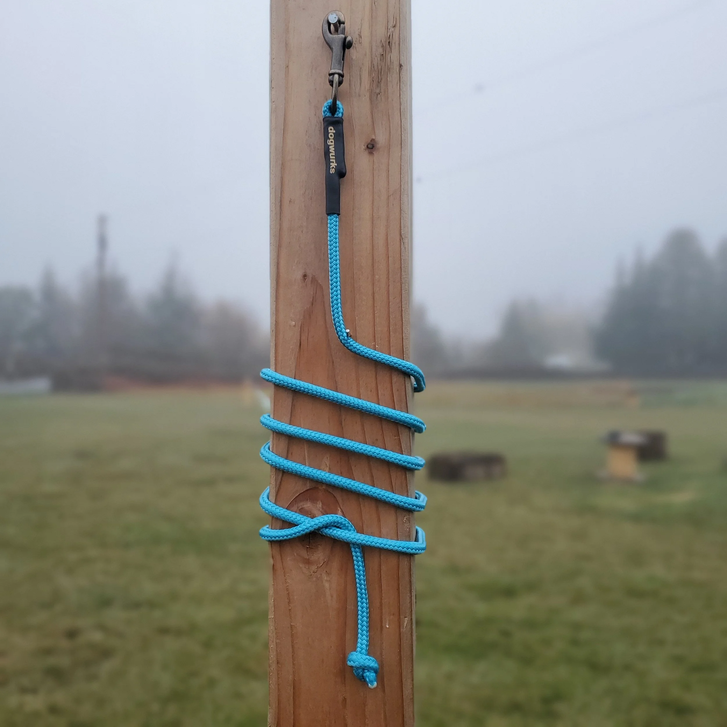 Blue climbing rope tied around a vertical wooden post, with a metal hook at the top, outdoors with a grassy field and cloudy sky in the background.