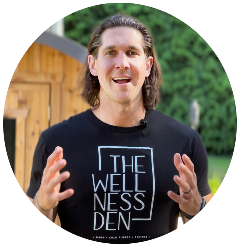 A man with long brown hair wearing a black T-shirt with 'The Wellness Den' written on it, standing outdoors with a wooden structure and green foliage in the background, speaking and gesturing with his hands.