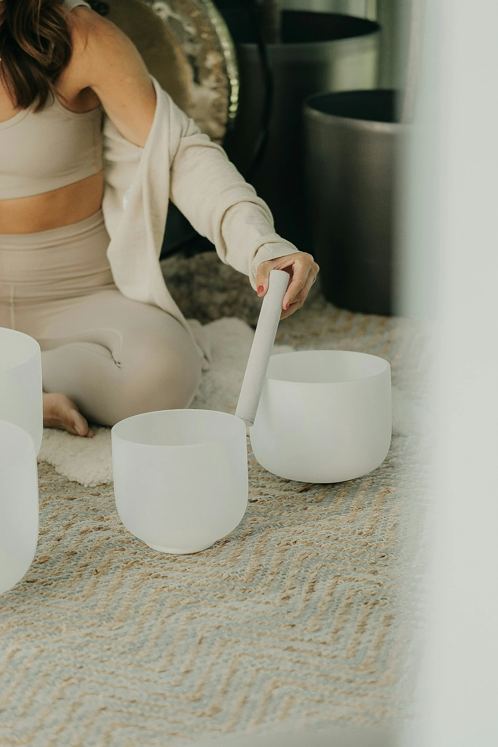 A woman sitting on a beige rug, dressed in neutral-colored workout clothes, playing crystal singing bowls with a wooden mallet. In the background, there are black and white cylindrical planters.