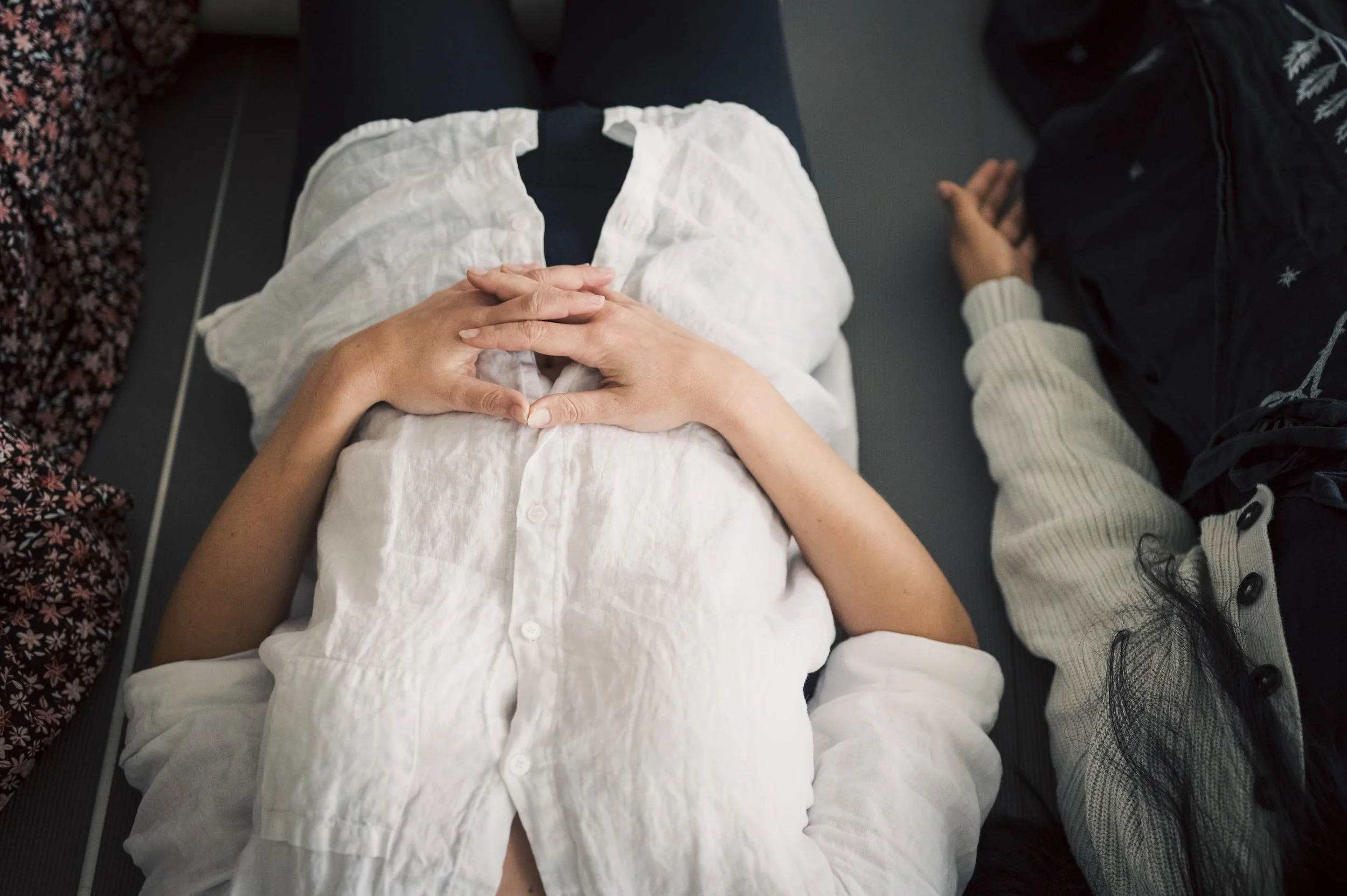 View from above of two people sitting on benches, one with hands folded on lap, the other with hands to sides, wearing a white blouse, dark pants, and a dark jacket, with a focus on their hands and laps.