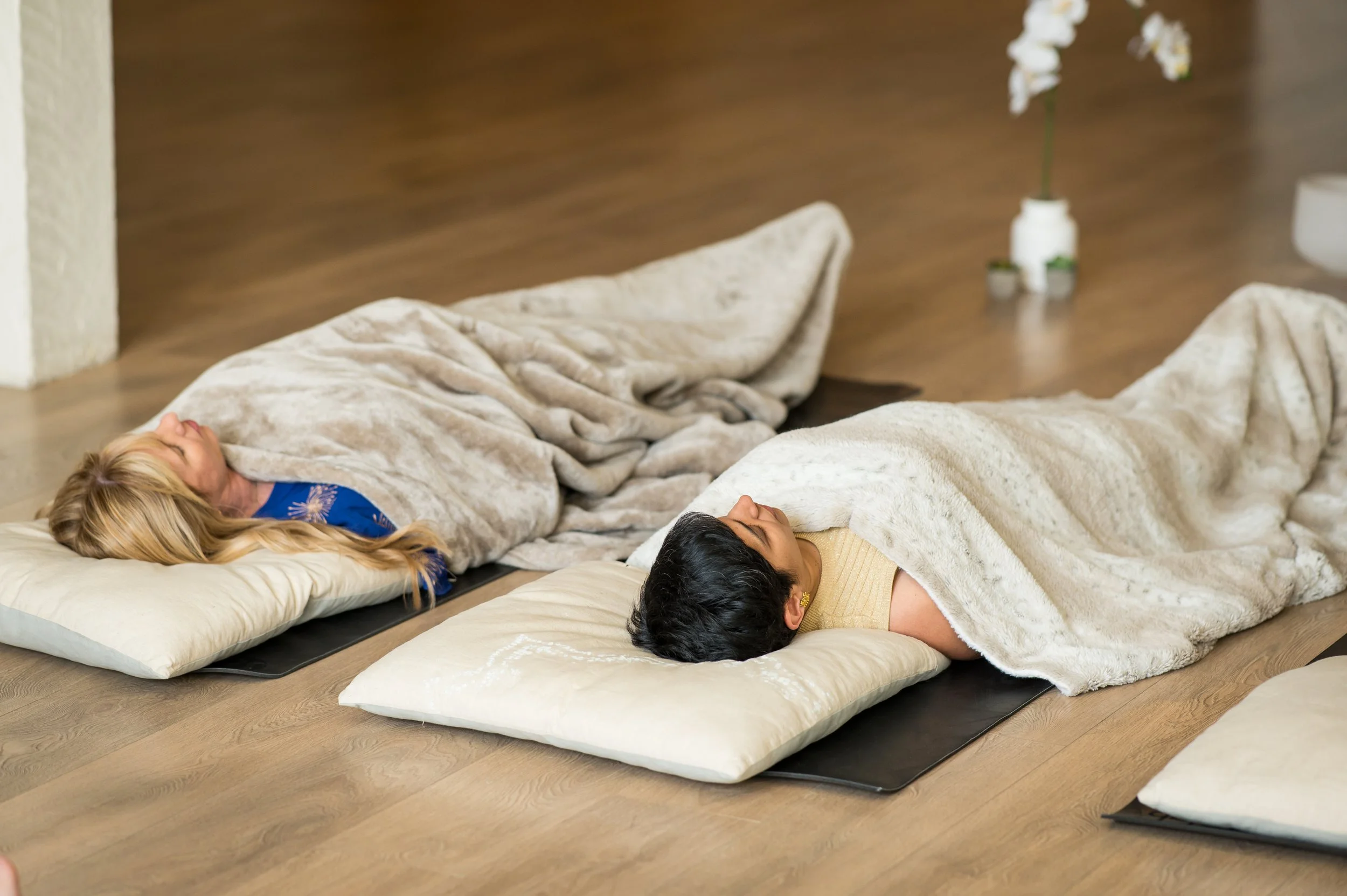 Three people lying on cushions on a wooden floor, covered with a beige blanket, in a room with minimal decor and a white vase with white flowers in the background.