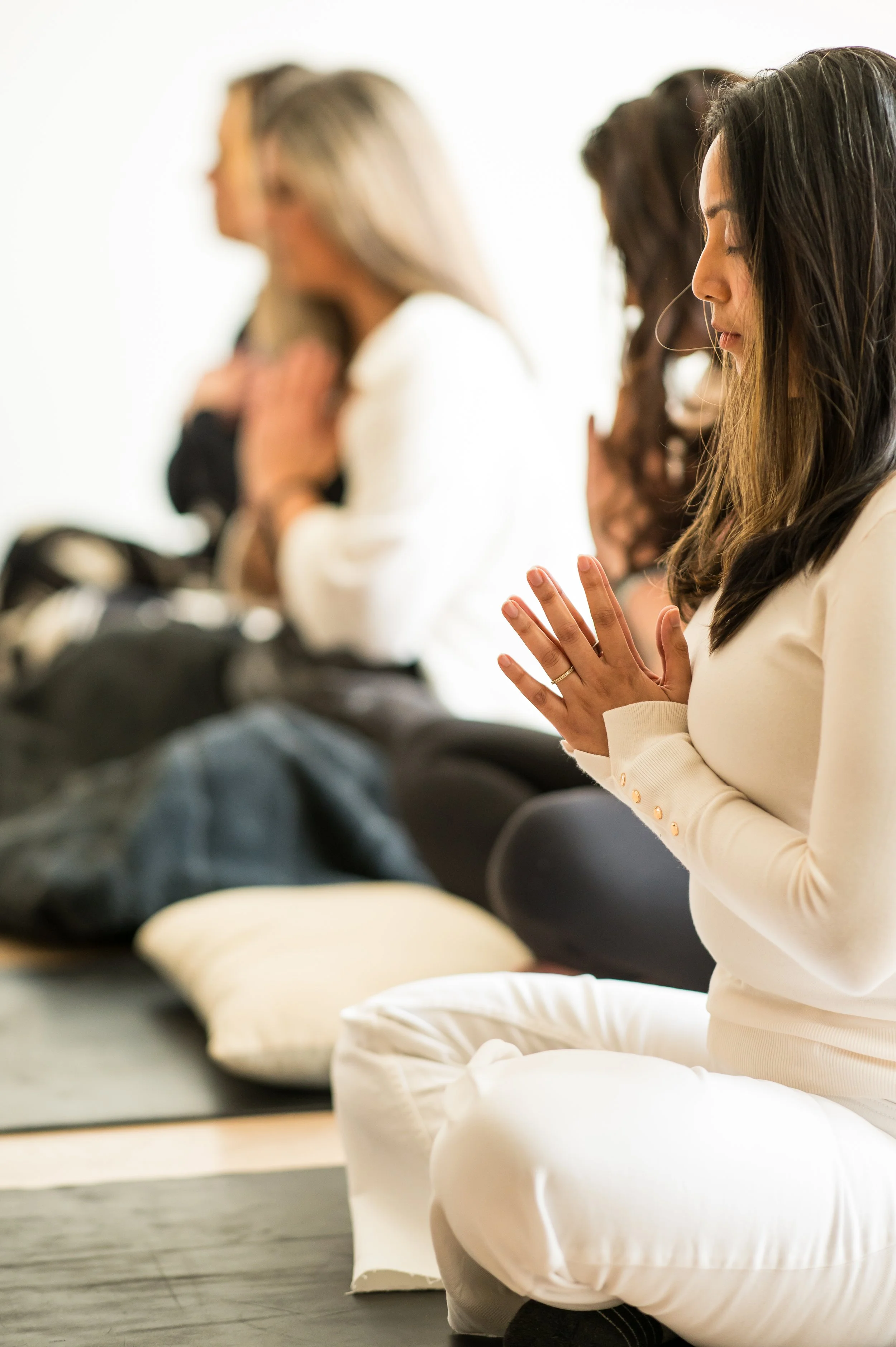 Women engaged in meditation or prayer, seated on cushions with hands in prayer position, in a peaceful indoor setting.