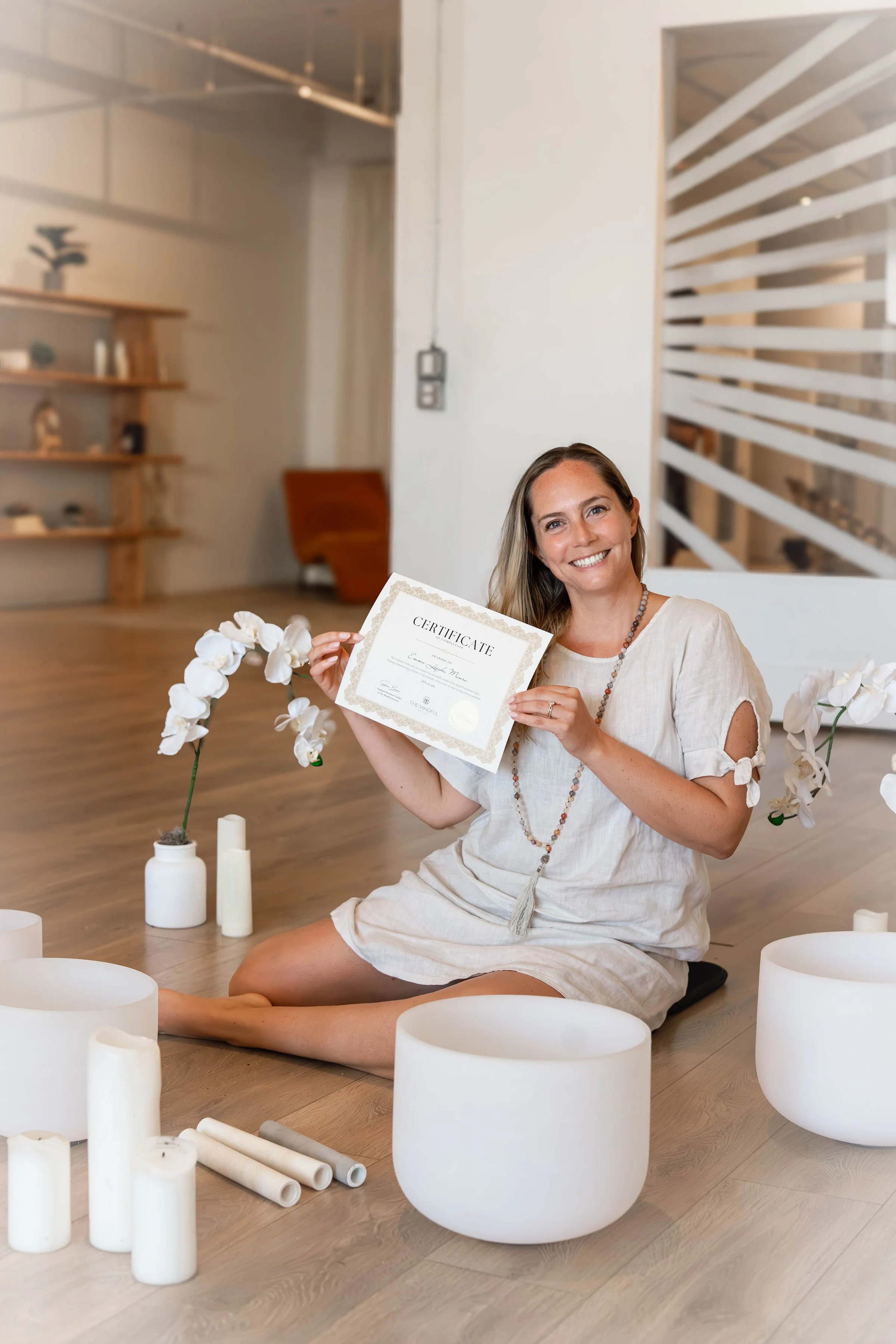 A woman sitting on a wooden floor, holding a certificate and smiling. She is surrounded by white candles, white ceramic bowls, and white orchids in vases. The setting appears to be a modern, minimalistic room with wooden shelves and a partially visible staircase in the background.