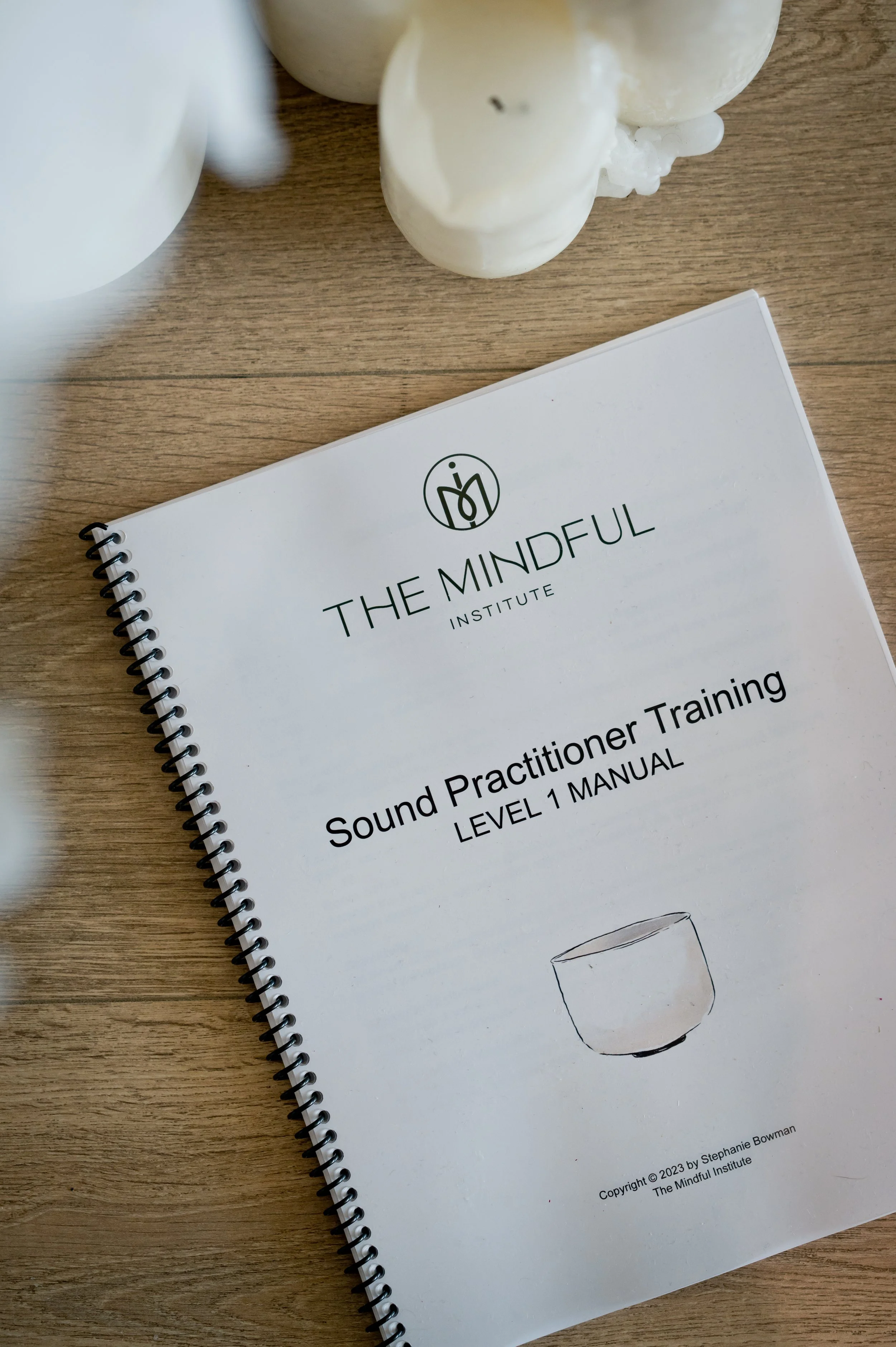 Cover of The Mindful Institute Sound Practitioner Training manual on a wooden table with white candles nearby.