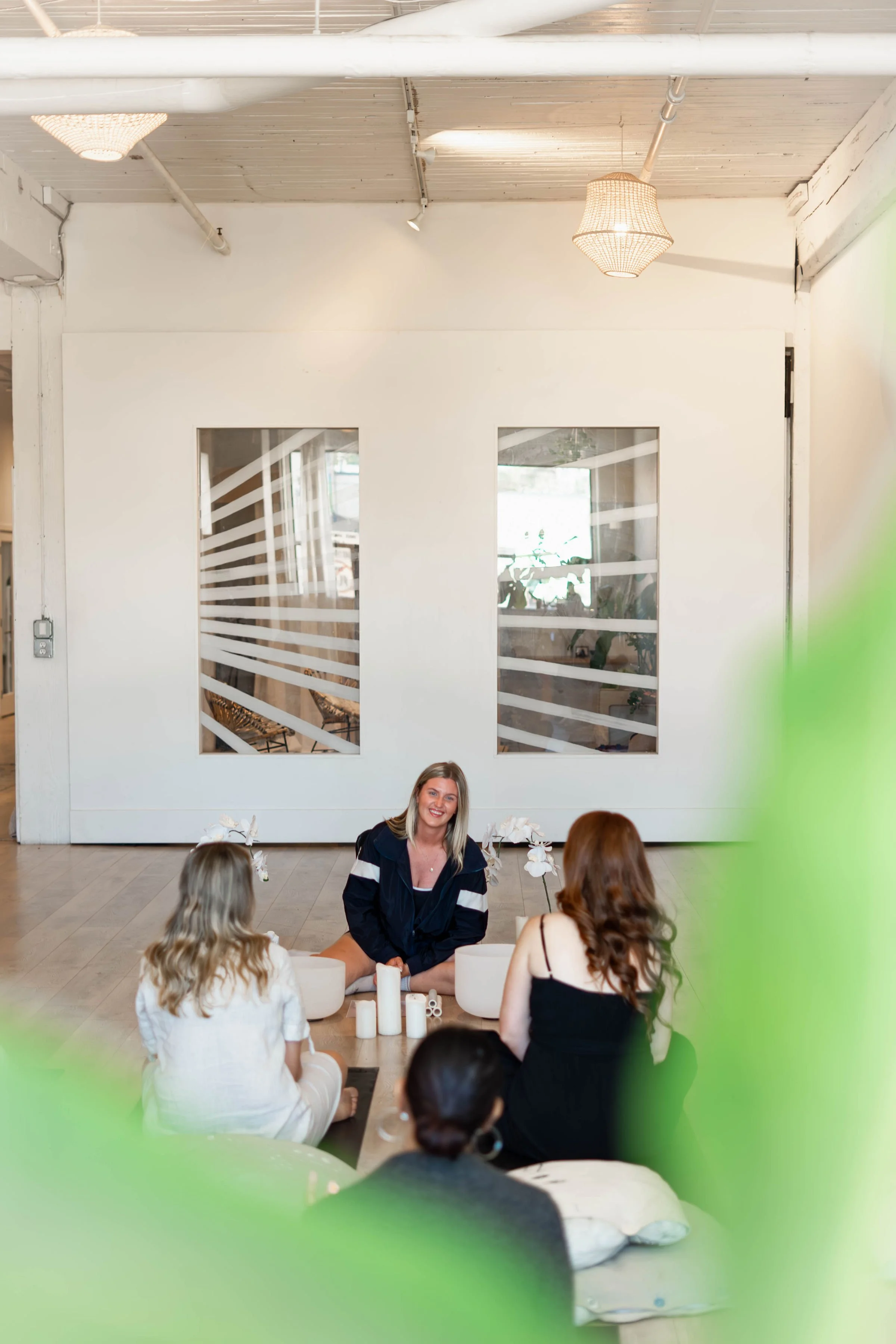 A woman leading a meditation session with four women seated on cushions in a room with white walls, wooden floors, and decorative lighting, seen through a partial green plant in the foreground.
