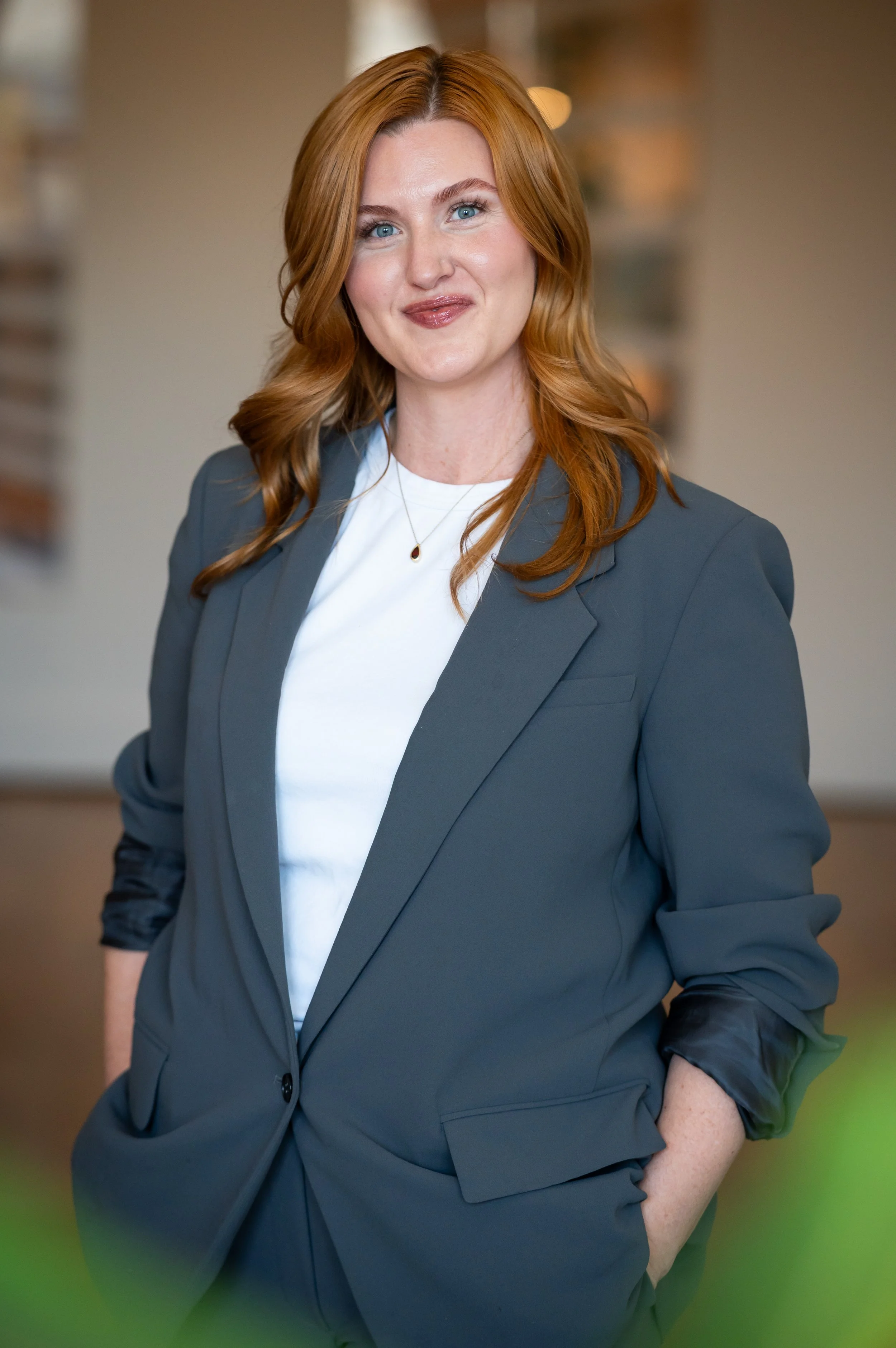 A woman with long red hair, blue eyes, and wearing a gray blazer over a white shirt, standing indoors with a blurred background.