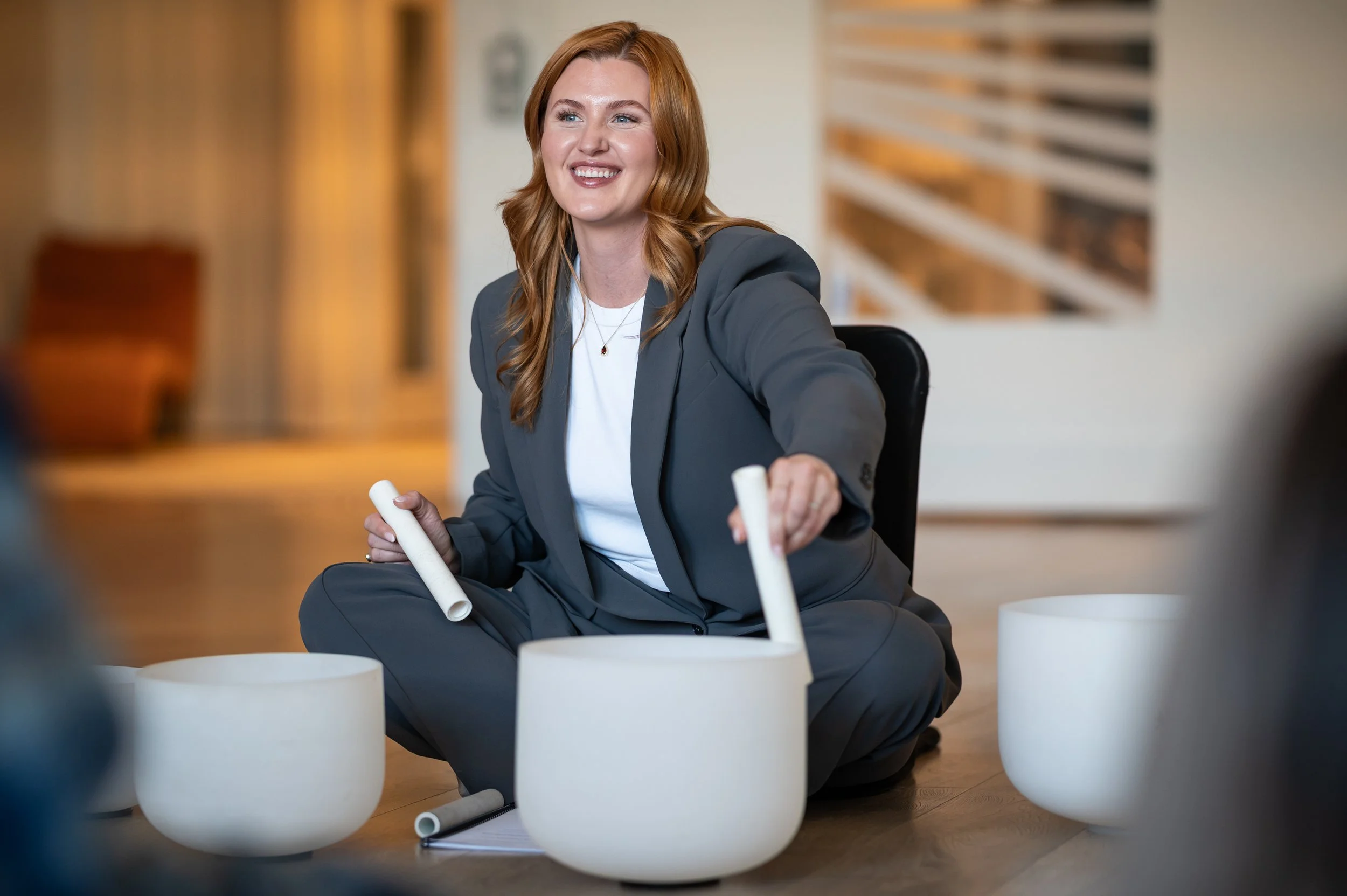A woman with red hair, wearing a gray suit, sitting on the floor with white singing bowls around her, holding two white mallets.