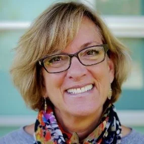Headshot of Penny Carver wearing dark-rimmed eyeglasses and a vibrant colorful scarf.