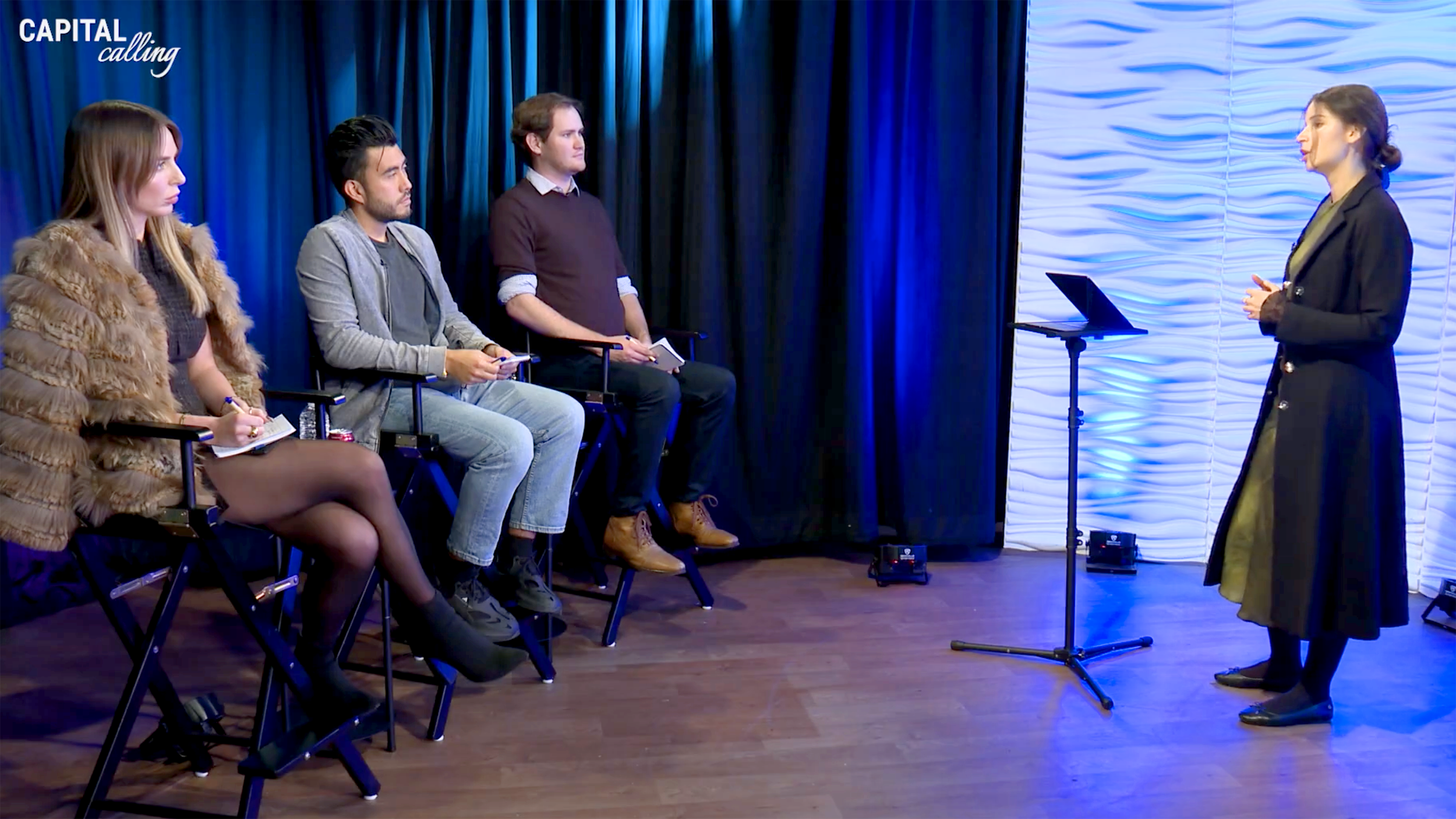 3 Judges sitting on the left of image listening to a presentation by a young woman on the right side of the image. A curtain slit with vibrant colors is behind the judges, and vibrant wavy panels are behind the presenter.