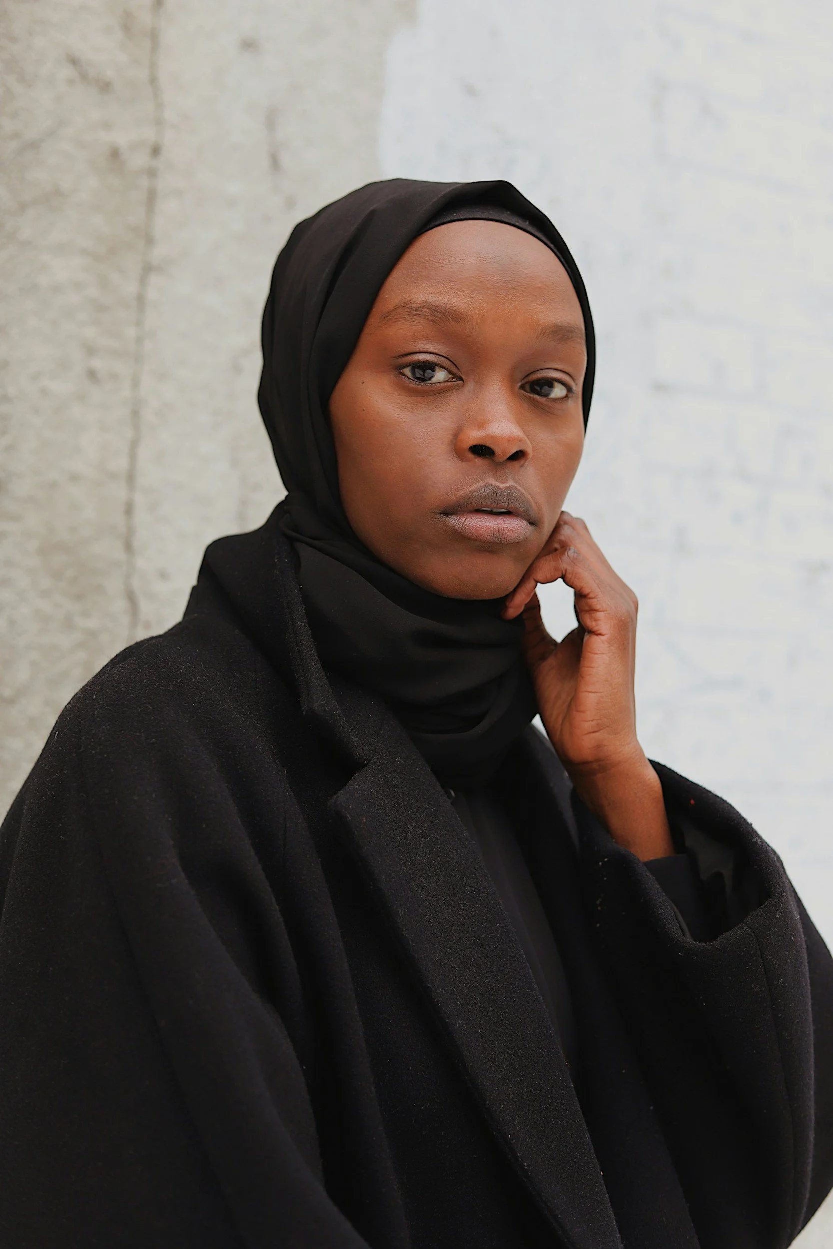 Close-up of a woman wearing a black headscarf and coat, standing against a textured wall.