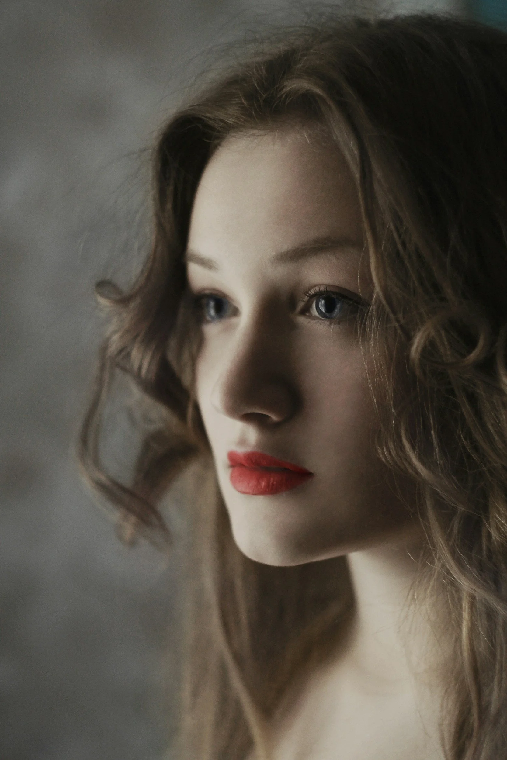 A close-up portrait of a woman with wavy brown hair, blue eyes, and red lipstick, gazing thoughtfully.