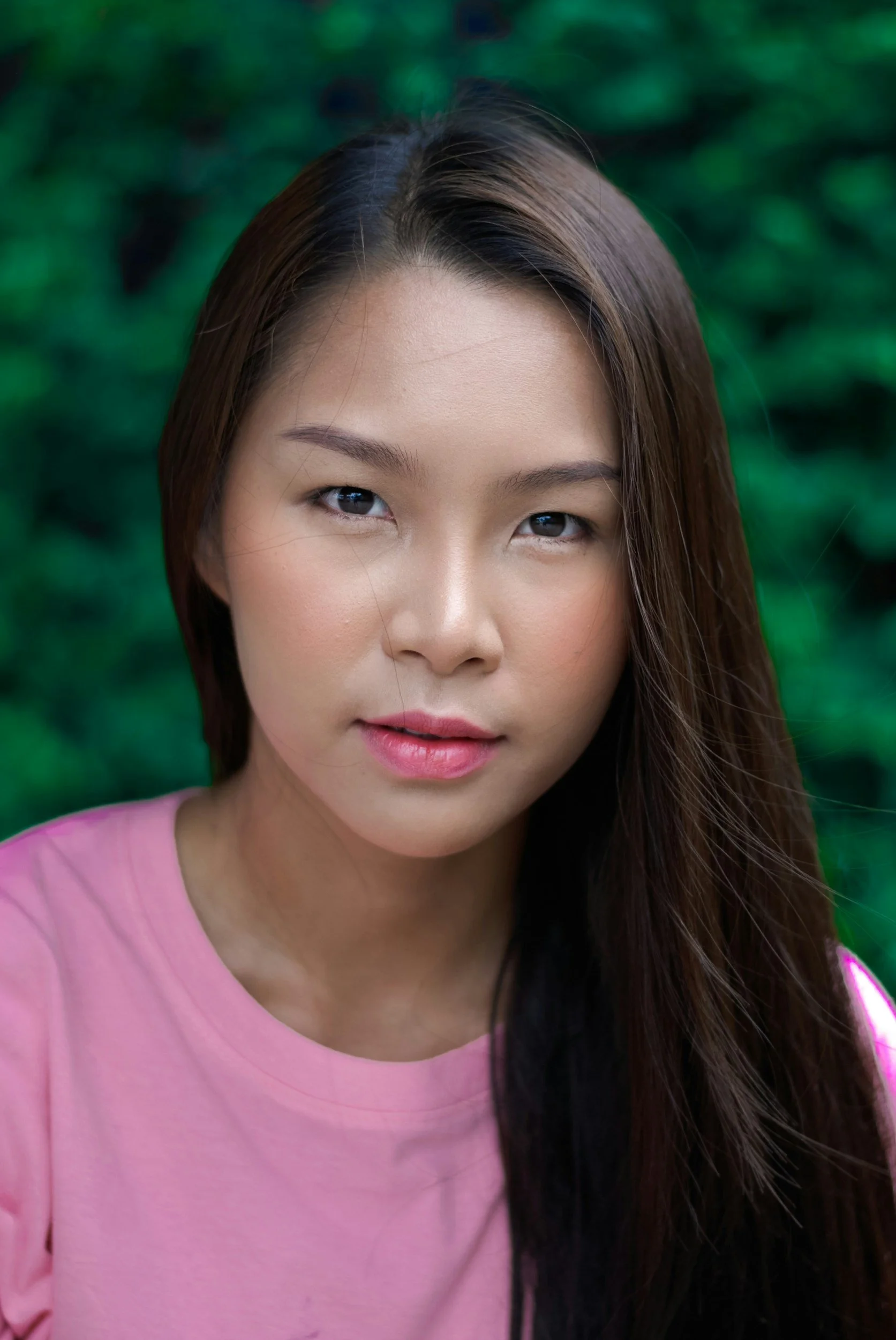 Close-up portrait of a young woman with long dark hair, wearing a pink shirt, in front of a green outdoor background.
