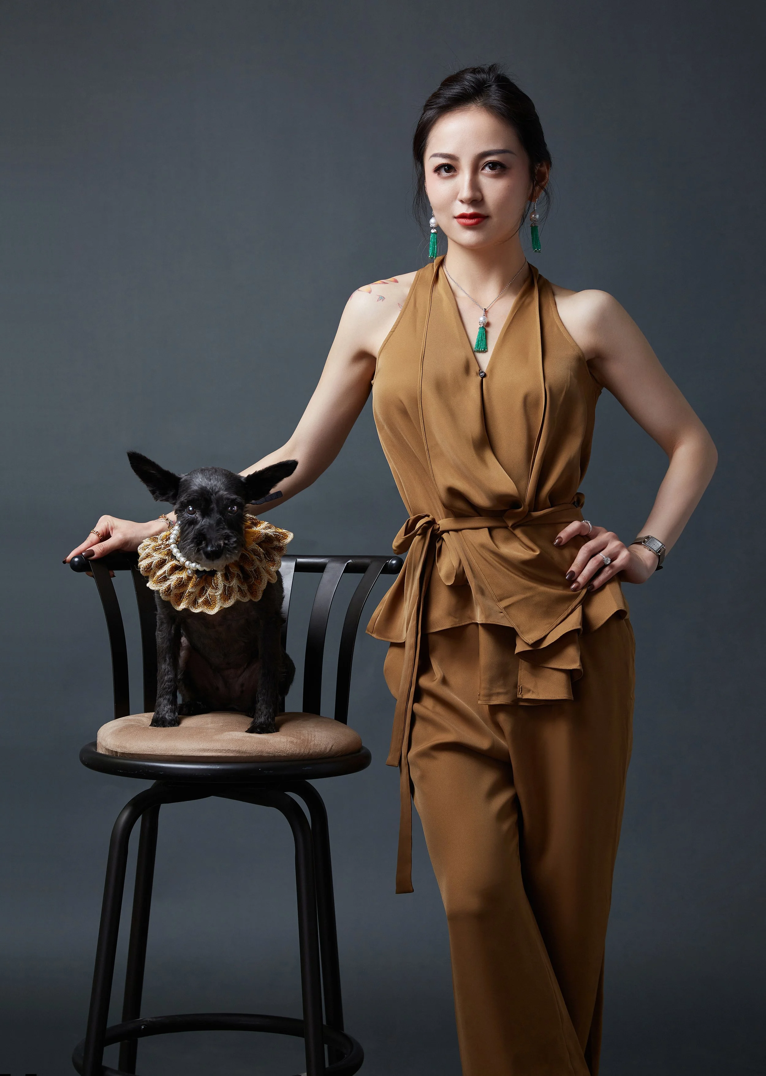 A woman in a brown sleeveless jumpsuit with a waist tie poses with a black dog sitting on a beige cushioned chair. The dog is wearing a ruffled collar. The woman has dark hair styled back and is accessorized with green tassel earrings, a matching necklace, and a watch. She is standing against a dark gray background.