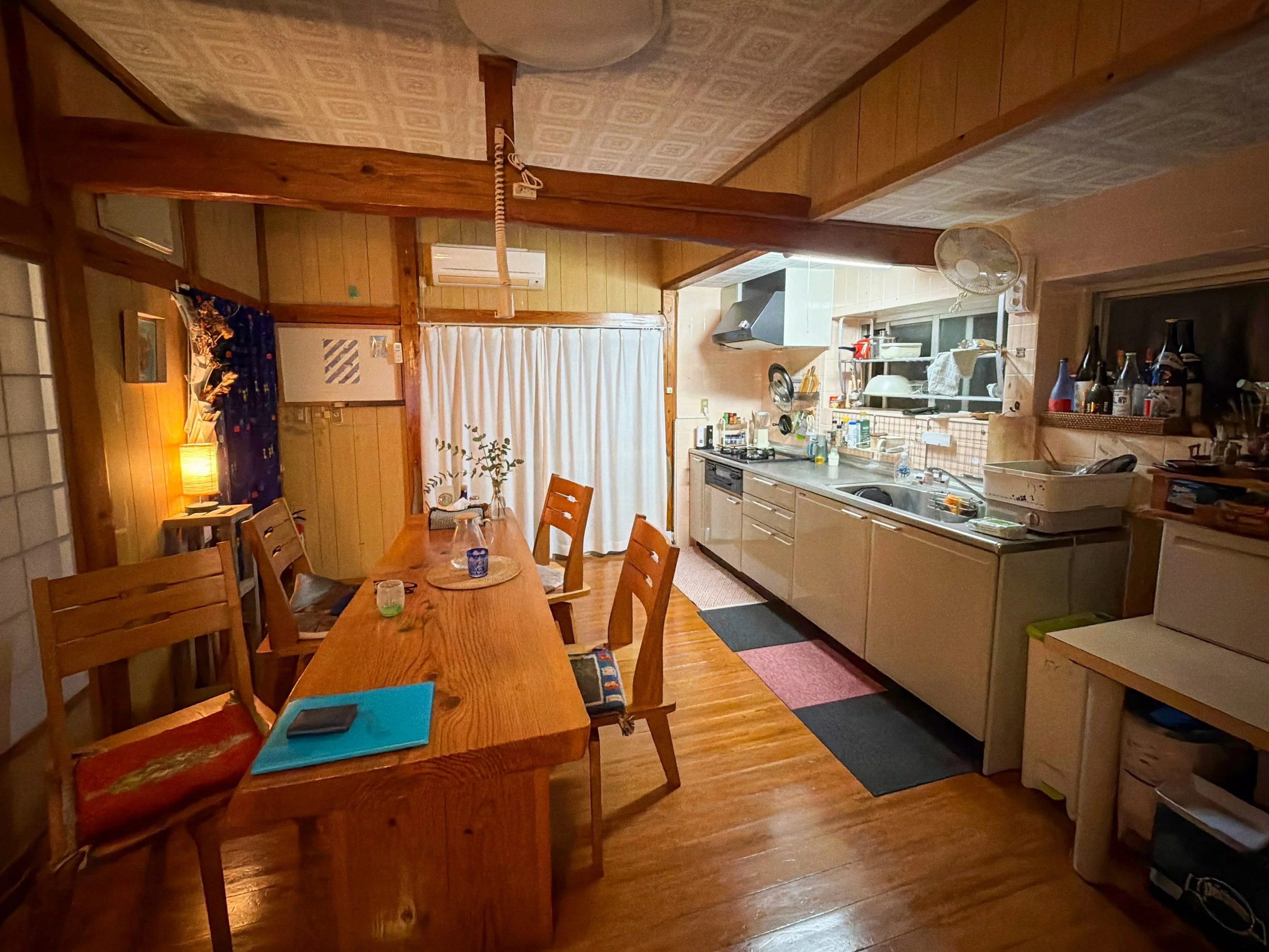 木の温もりを感じる、昔ながらの和風住宅の居間です。中央には木製の大きな長方形の食卓があり、周囲に数脚の椅子が並んでいます。右側には食器や調理器具が並ぶキッチンがあり、奥には白いカーテンがあります。暖色の照明が部屋全体を柔らかく照らし、落ち着いた家庭的な雰囲気を醸し出しています。