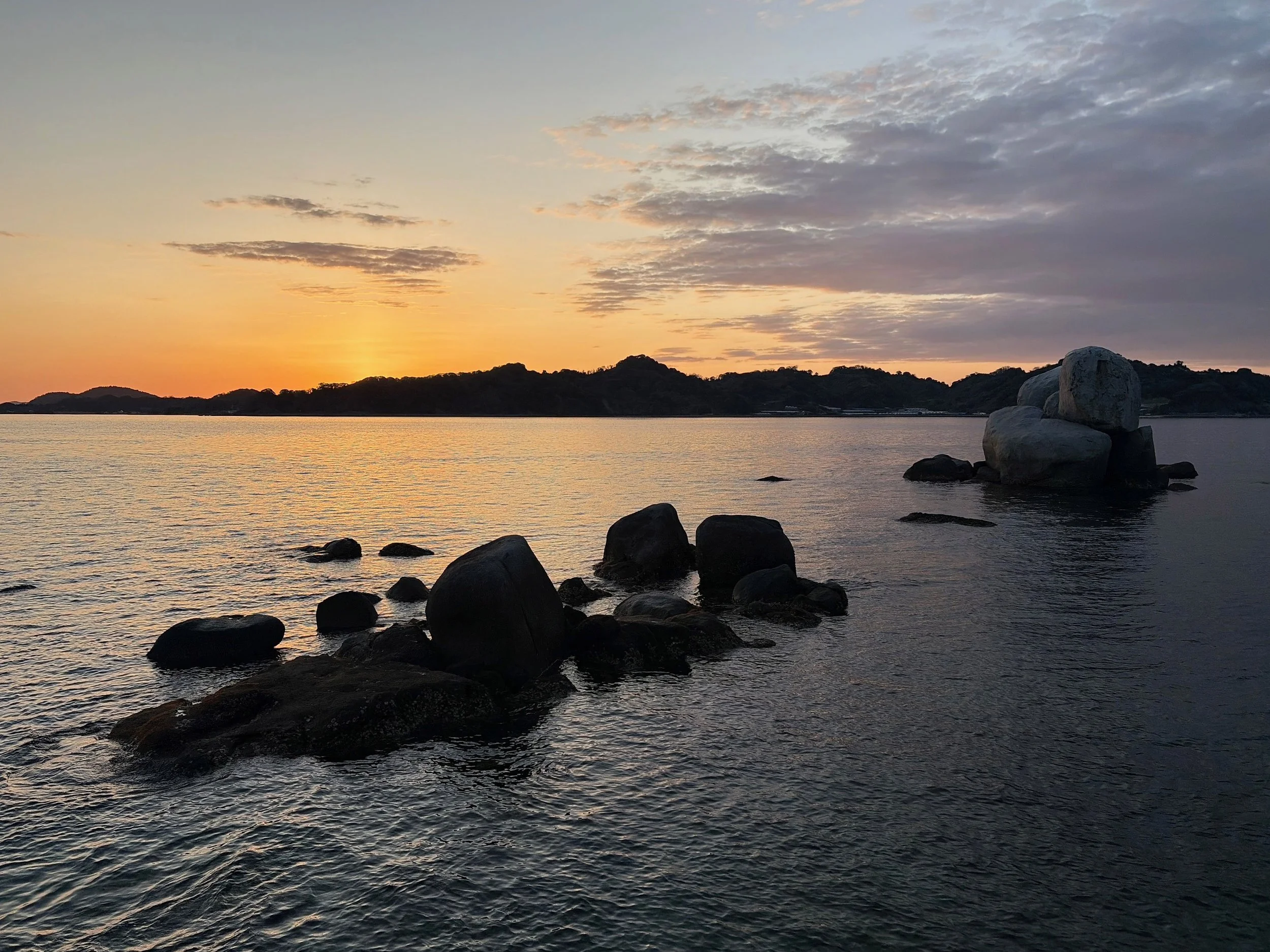 夕暮れの海を写した風景です。水平線の左側では太陽が沈み、空と海を淡いオレンジ色に染めています。手前の海には大小さまざまな黒い岩が連なり、穏やかな波が岩の間を静かに流れています。右奥には、丸い大きな岩が積み重なった特徴的な岩礁があります。遠くには低い山々が黒いシルエットで横一列に広がり、上空には厚い雲が夕日に照らされています。