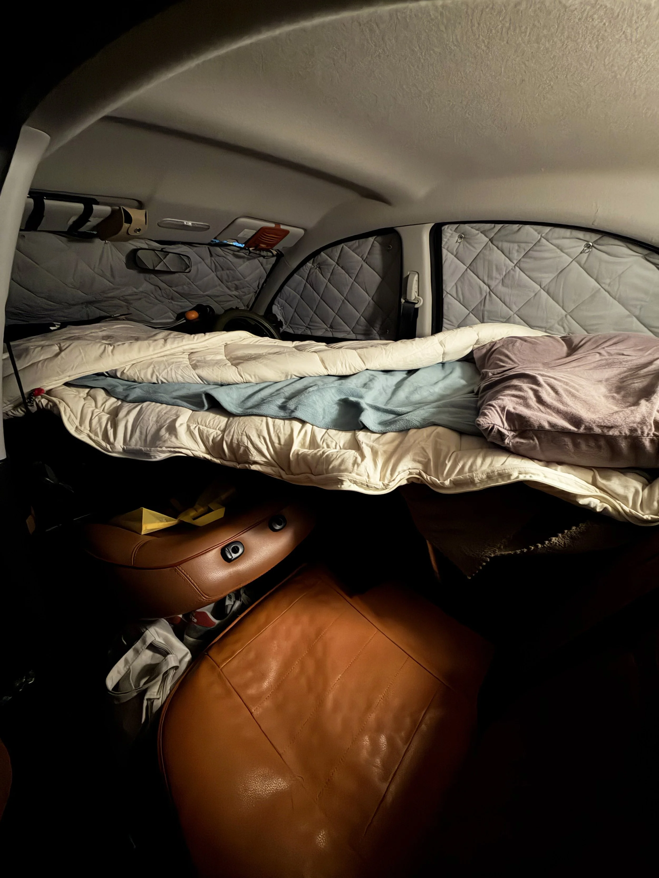 車の後部座席をベッドとして使えるように整えた車内の写真です。座席を倒した広いスペースには、白い掛け布団と水色のブランケットが敷かれ、右側には薄紫色の枕があります。窓には銀色のキルティング製の目隠しが取り付けられています。手前には茶色の革張りの座席があり、限られた車内空間を工夫して、快適に眠れる場所を作っている様子が伝わります。