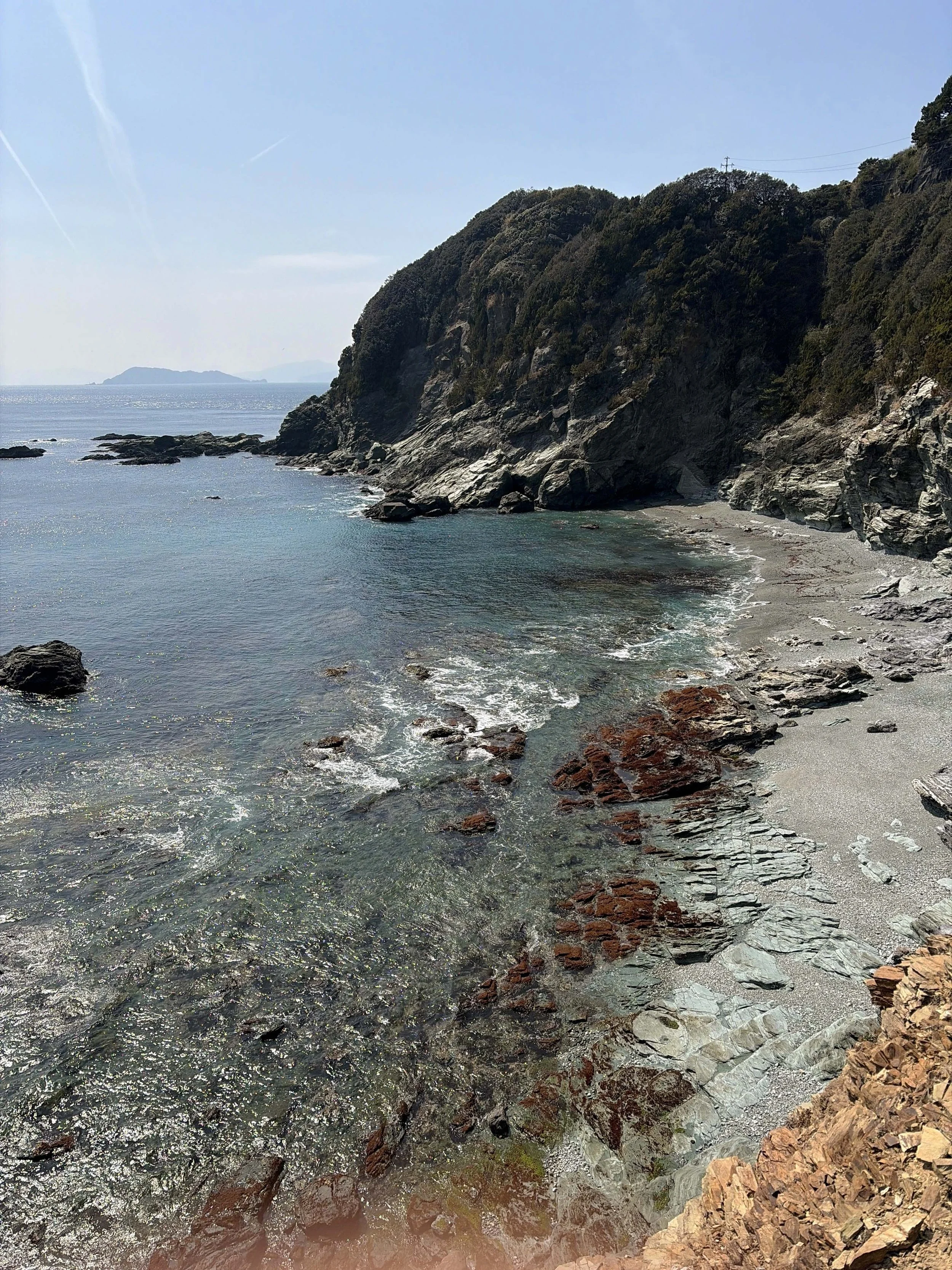 青い海に面した岩場の海岸を写した風景です。手前から沖へ向かって透明感のある海が広がり、太陽の光を受けて水面がきらきら輝いています。右側には灰色や茶色の岩が重なる険しい海岸線と、木々に覆われた大きな崖があります。波が岩場に静かに打ち寄せ、遠くの水平線には島影がぼんやりと浮かんでいます。晴れた空と穏やかな海が印象的な、自然豊かな海辺の景色です。