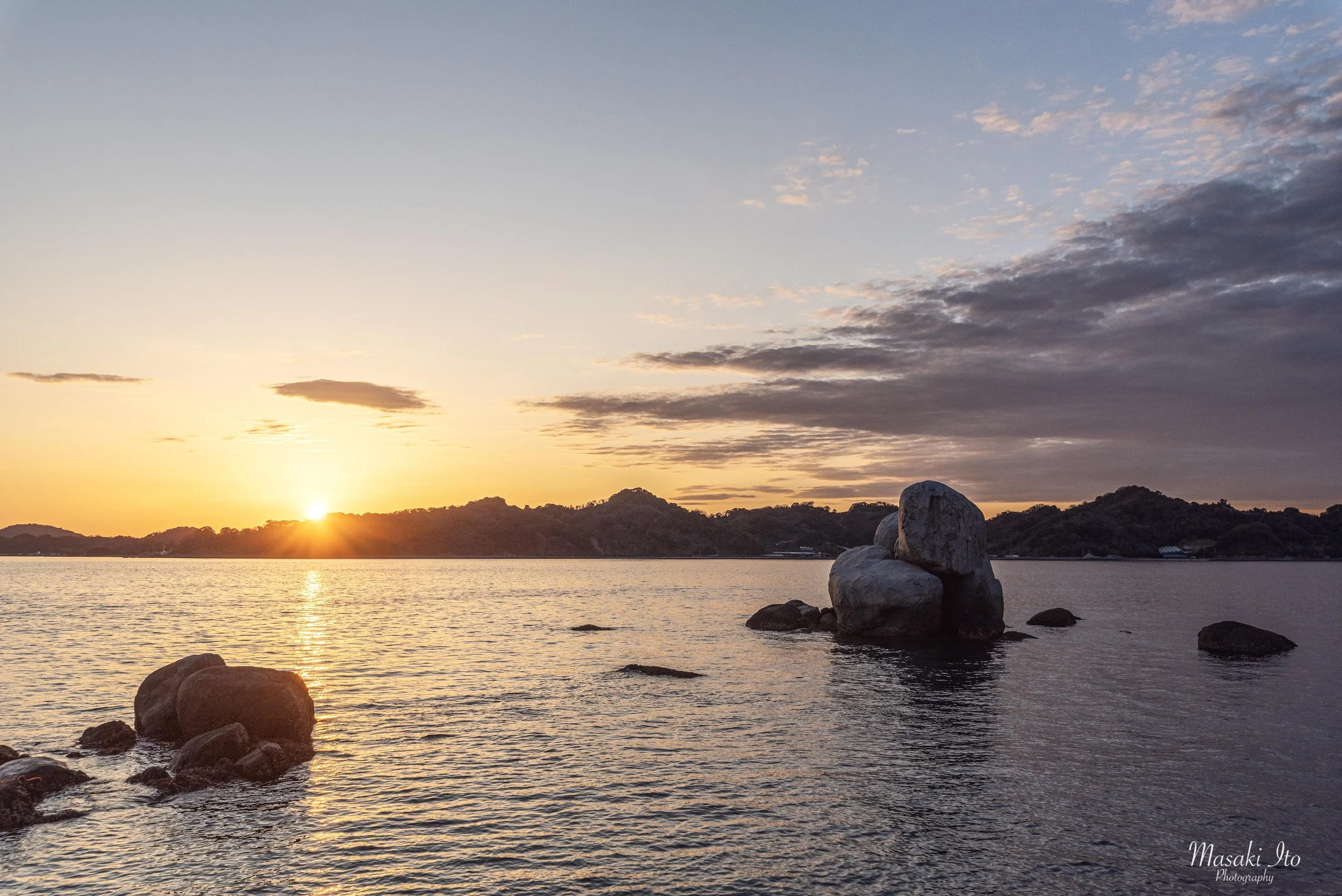 夕暮れの海を撮影した美しい風景です。水平線の左側に太陽が沈みかけ、海面には黄金色の光が一直線に伸びています。中央右には、大小の丸い岩が積み重なったような大きな岩礁が海から突き出し、周囲にも岩が点在しています。遠くには低い山々が黒いシルエットで連なっています。空は夕日の橙色から淡い青色へ変化し、右上には厚い雲が広がっています。