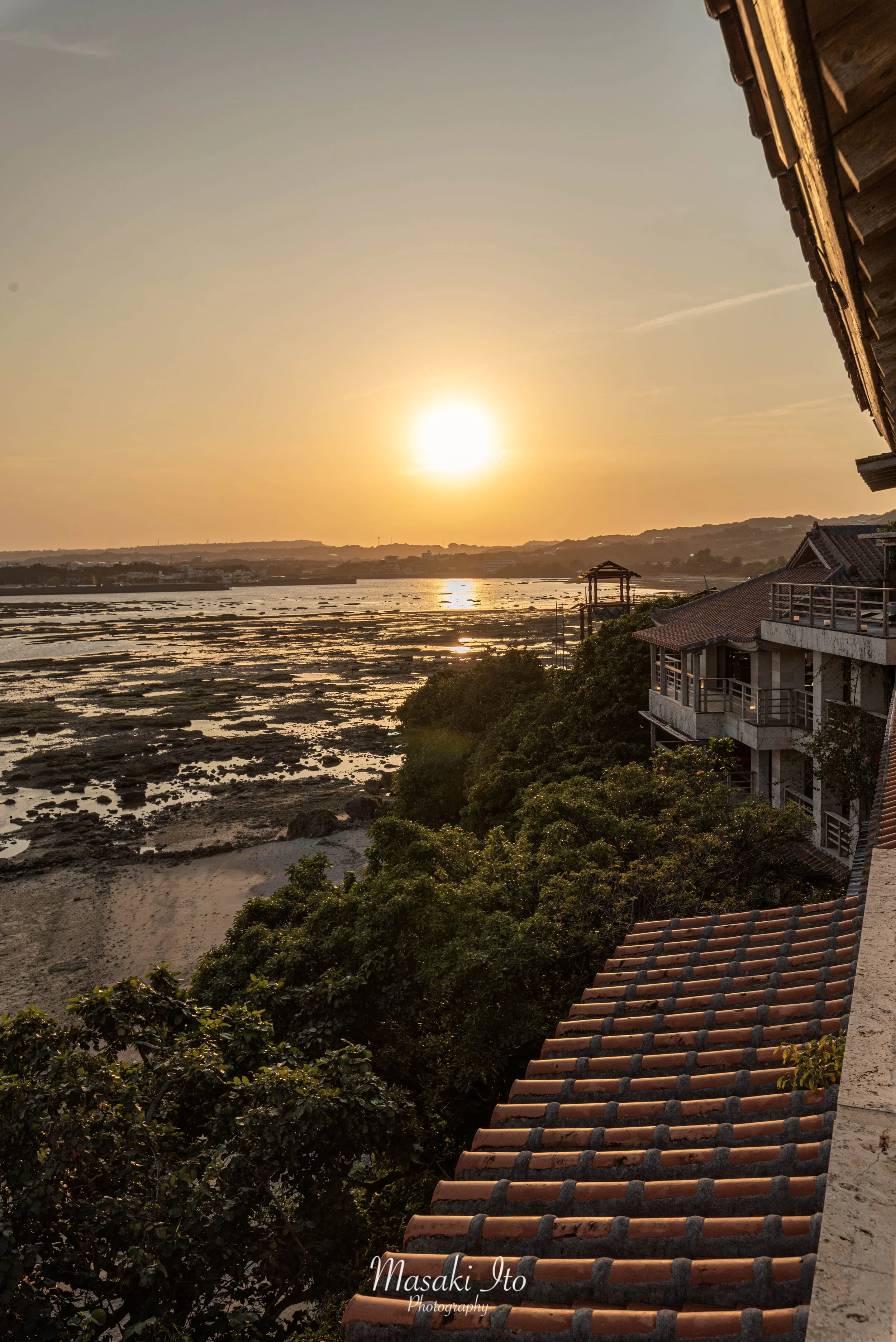 沖縄県南城市にあるホテル百名伽藍から望む、美しい夕景を撮影した写真。水平線近くに広がる海と、岩礁が点在する浅瀬に夕日のオレンジ色の光が反射している。右側には赤瓦屋根の建物が並び、手前には緑豊かな木々が広がる。空を黄金色に染める太陽と、沖縄らしい海岸風景、伝統的な屋根の質感が調和した、温かく落ち着いた雰囲気の一枚。