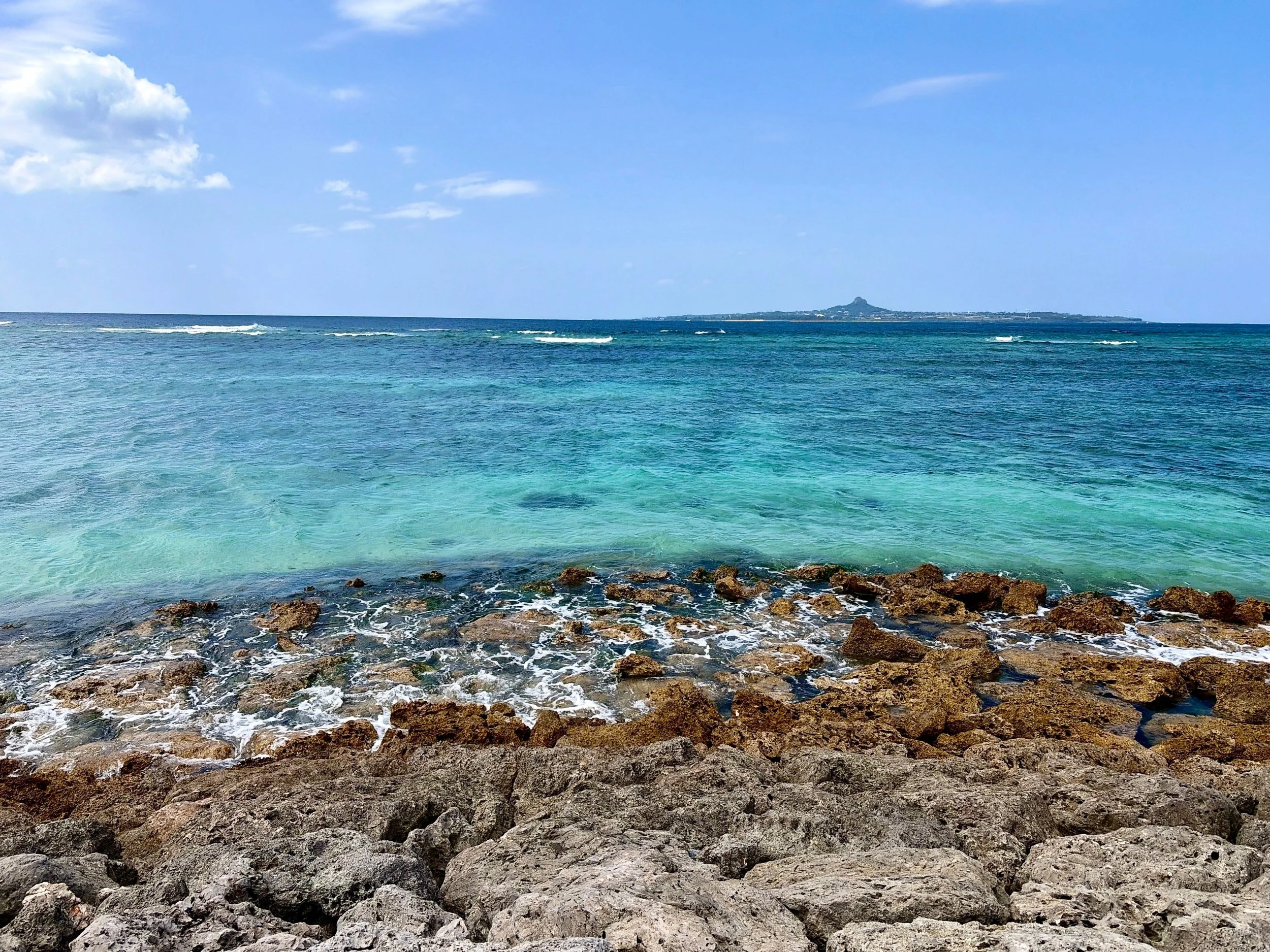 青く澄んだ空の下、広大な海が一面に広がっています。手前には、茶色や灰色のごつごつした岩場があり、白い波が岩の間を縫うように寄せています。海は浅瀬から沖に向かって、透明感のあるエメラルドグリーンから深い青へと変化。水平線の先には細長い島が浮かび、その中央付近には小さな山が見えます。爽やかで開放感のある南国の海辺です。