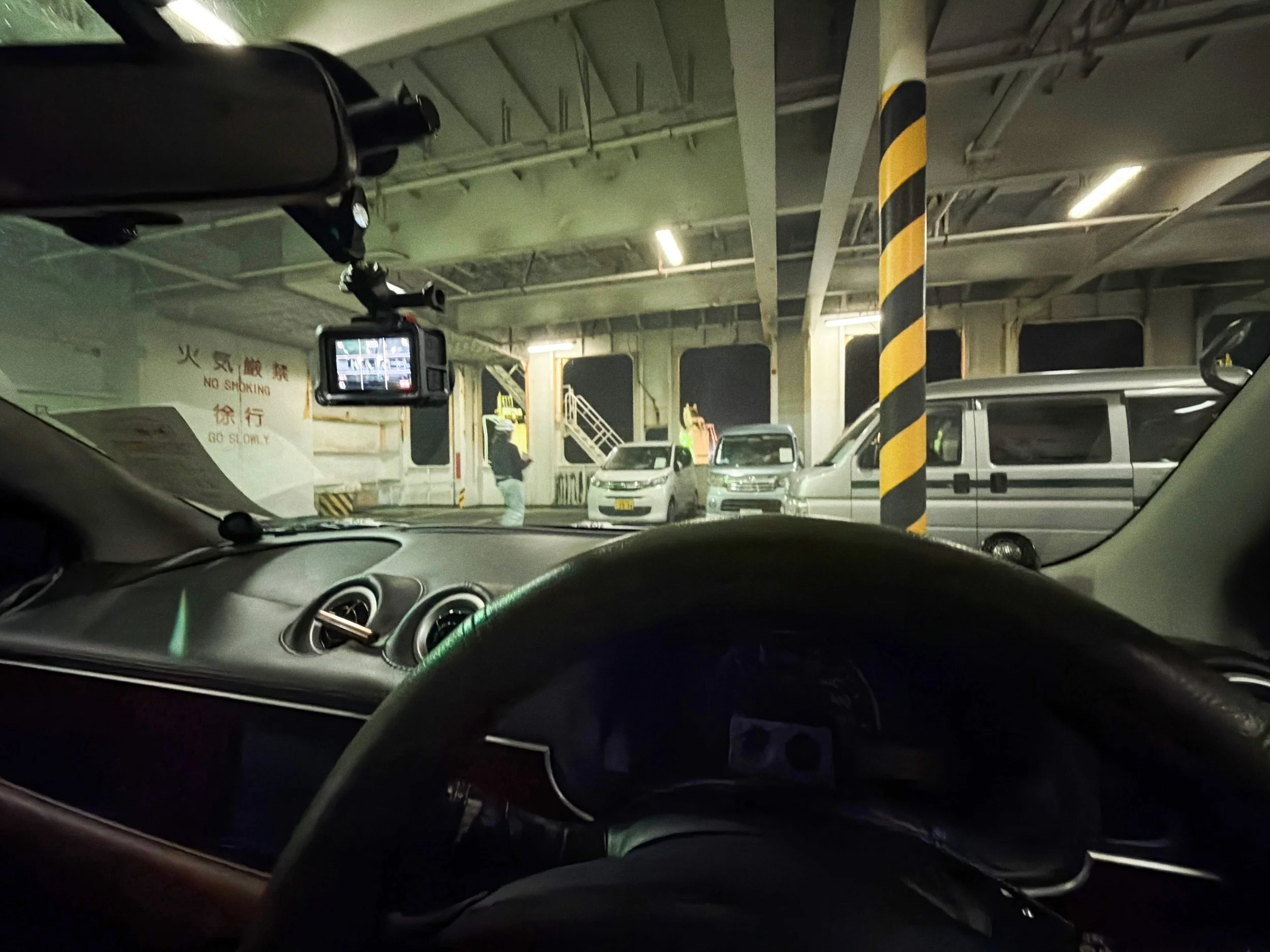 車の運転席から、フェリーの車両甲板を見渡した写真です。手前には大きなハンドルとダッシュボードがあり、フロントガラス越しに駐車された車が並んでいます。中央には黄色と黒の斜め縞模様の柱が立ち、天井には白い照明や配管が見えます。奥には階段と作業する人の姿もあり、船内特有の無機質な雰囲気が漂っています。壁には「火気厳禁」「徐行」と日本語や英語、中国語の注意書きがあります。
