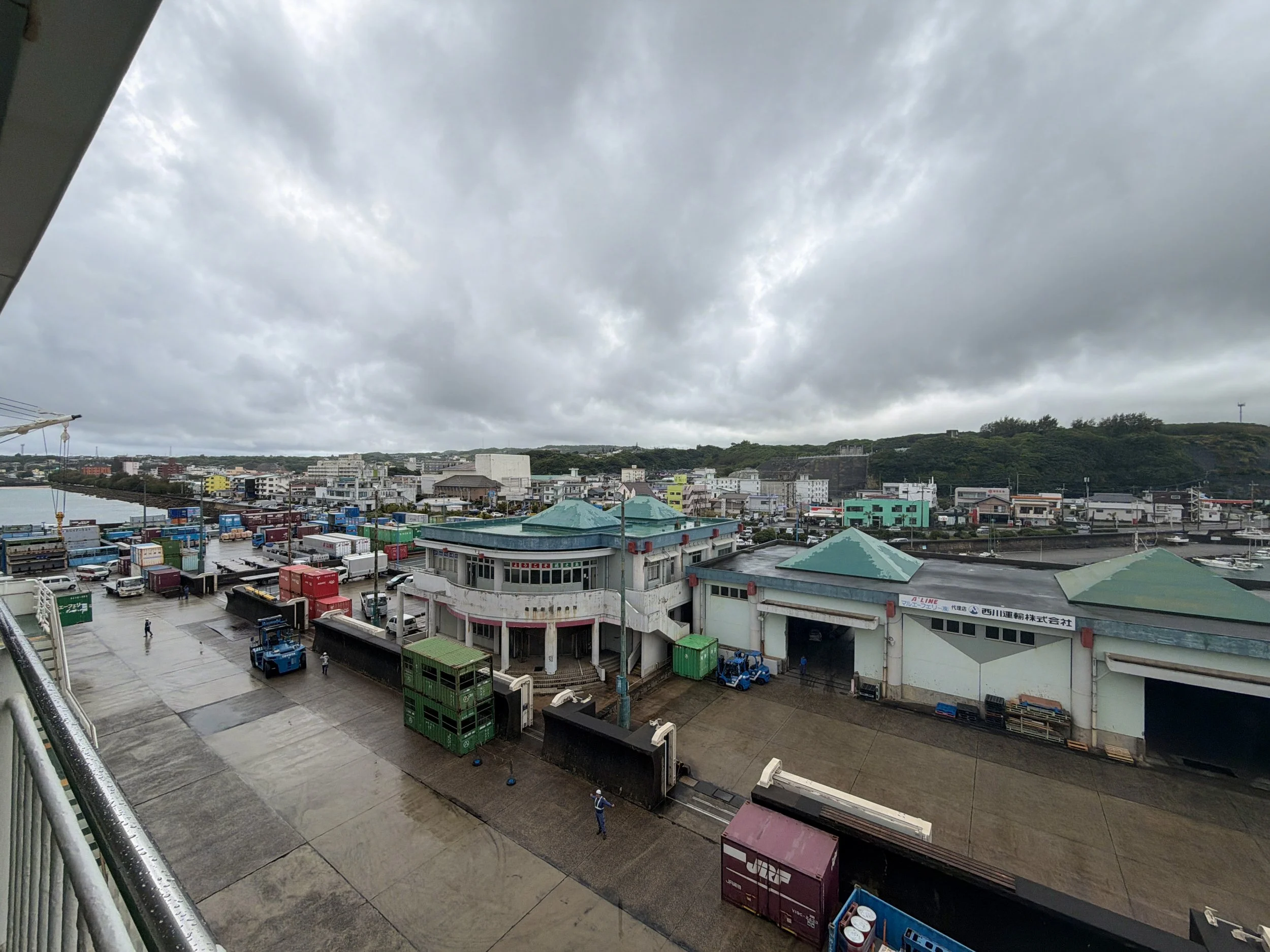 徳之島の亀徳港を高い場所から見下ろした風景です。雨に濡れた広い港の敷地には、赤や青、緑など色とりどりのコンテナが並び、貨物を運ぶ青いフォークリフトも見えます。中央には緑色の屋根を持つ港の建物があり、その奥には低い丘と亀徳の街並みが広がっています。空は厚い灰色の雲に覆われ、南の島らしい緑の丘との対比が印象的です。
