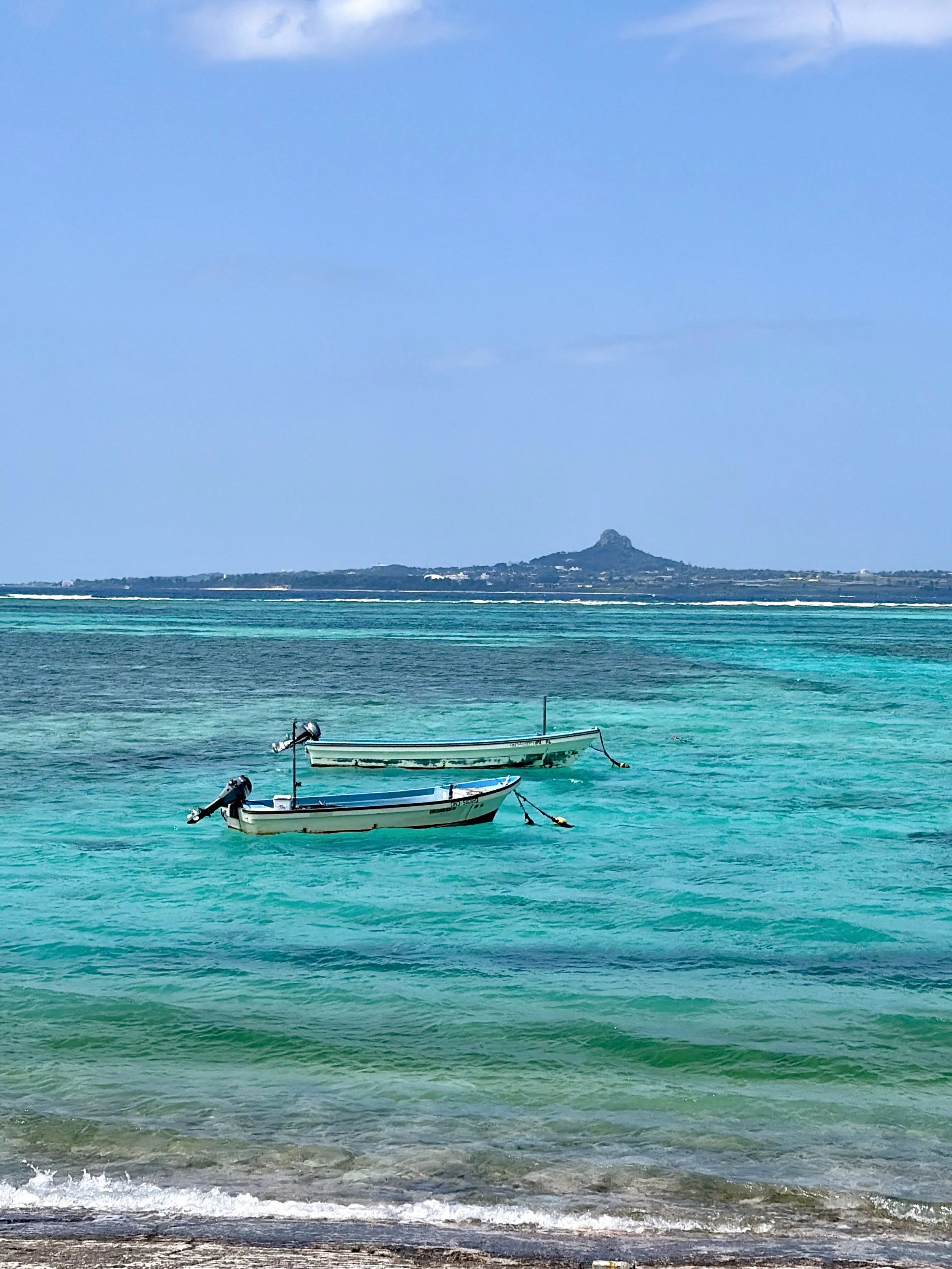備瀬の浜から見た、穏やかな海の風景です。手前には透明感のある浅瀬が広がり、エメラルドグリーンから青へと水の色が変化しています。波打ち際では白い波が静かに砕けています。海の中央付近には、小さな白いボートが2隻、ロープでつながれ浮かんでいます。遠くには細長い島影が横たわり、その中央に特徴的な尖った岩山が見えます。青空と海が一体となった、沖縄らしい開放的な景色です。