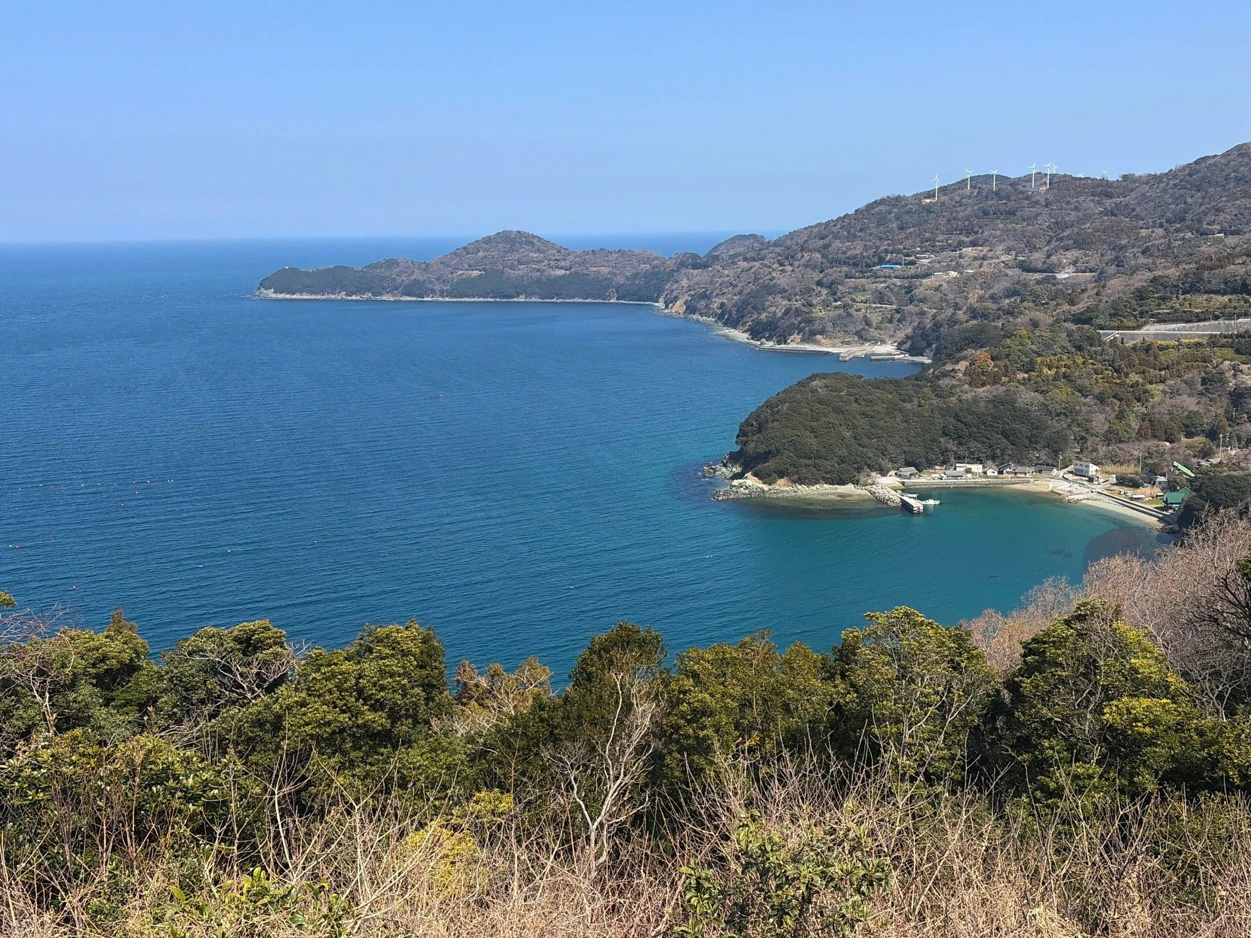 青空の下、眼下に広がる深い青色の海を望む高台からの風景です。海岸線は大きく湾曲し、手前には木々が生い茂っています。右側には小さな集落や港があり、山の斜面には道路や建物が見えます。遠くの山の尾根には白い風車が並び、穏やかな海と緑の山々が美しく調和しています。海は場所によって青緑色に変化し、透明感のある美しさを感じさせます。