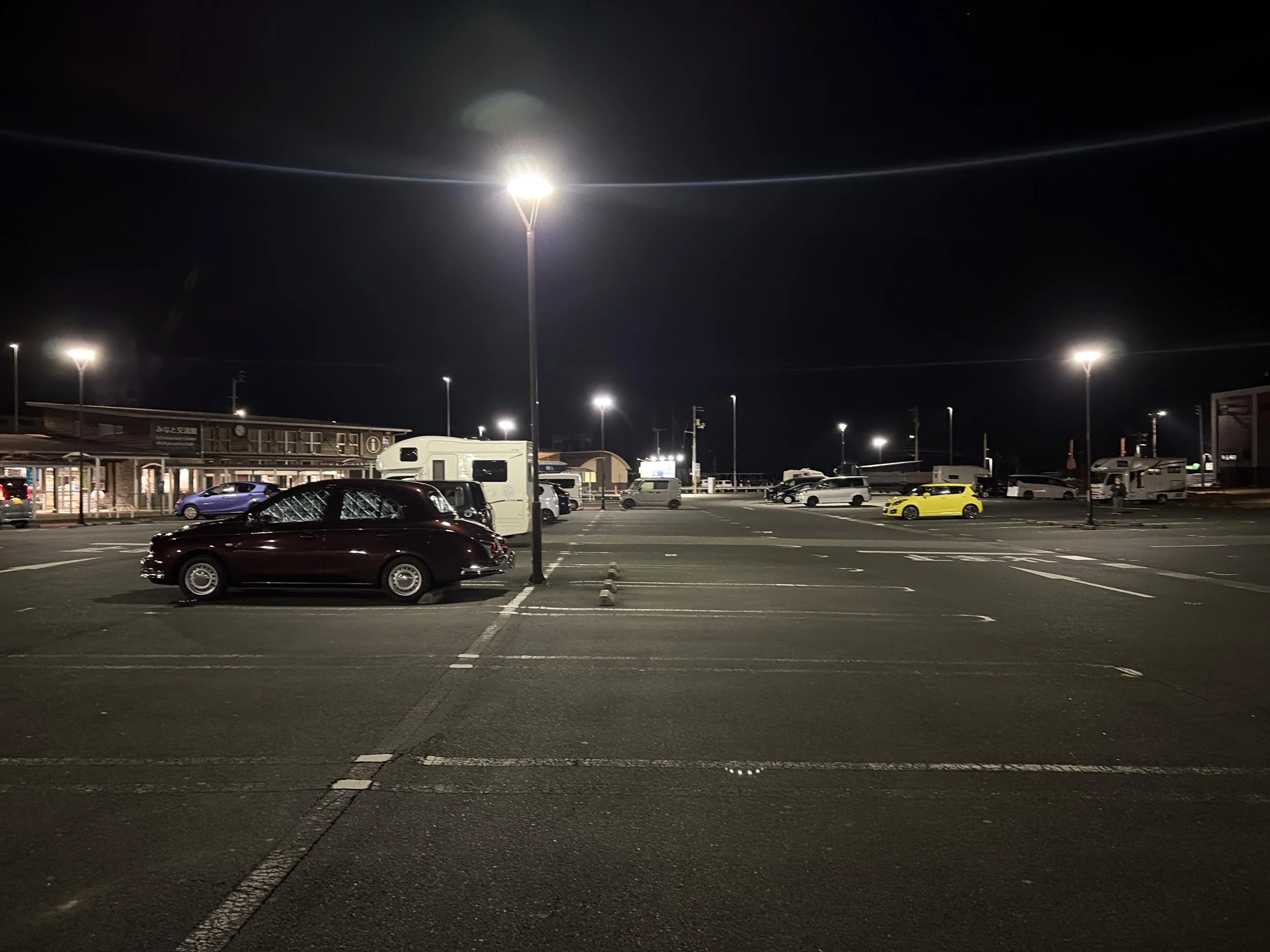 夜の広い駐車場を写した風景です。中央には白い街灯が高く立ち、周囲にも複数の街灯が並んで明るく駐車場を照らしています。手前にはワインレッドの乗用車が停まり、その奥にはキャンピングカーやさまざまな車が点在しています。右側には鮮やかな黄色い車も見えます。左奥には明かりのついた建物があり、黒い夜空との対比が印象的な、静かな夜の道の駅のような場所です。