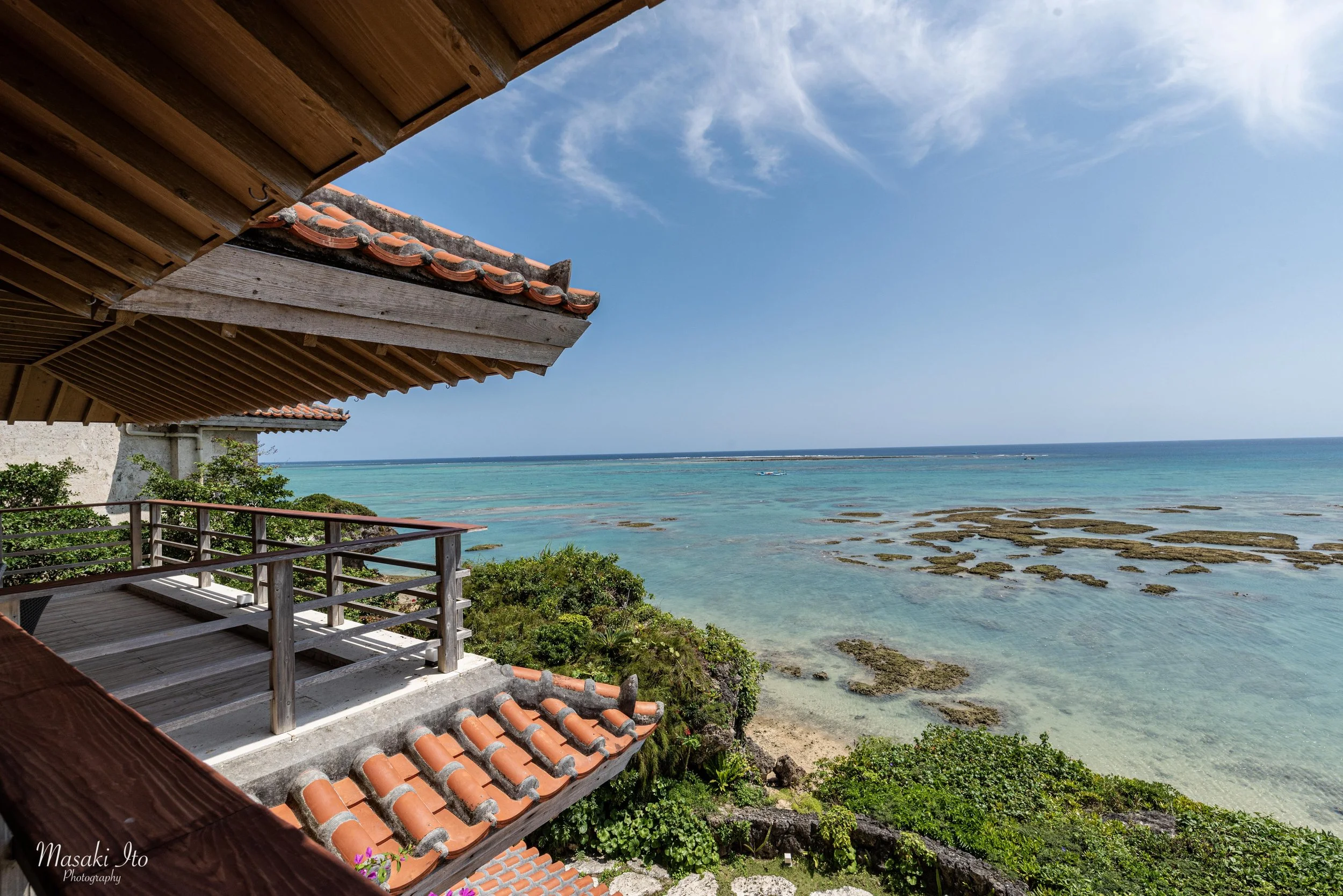 沖縄県南城市にあるホテル百名伽藍の海沿いの風景。写真の左側には、赤瓦屋根と木造の梁が特徴的な沖縄らしい建物があり、木製の手すりが付いたテラスから美しい海を望める。右側には、浅瀬のエメラルドグリーンの海が広がり、海面から顔を出す岩礁が点在している。遠くには水平線が伸び、青空には白い雲が流れる。伝統的な建築と南国の自然が調和した、開放感あふれる絶景である。