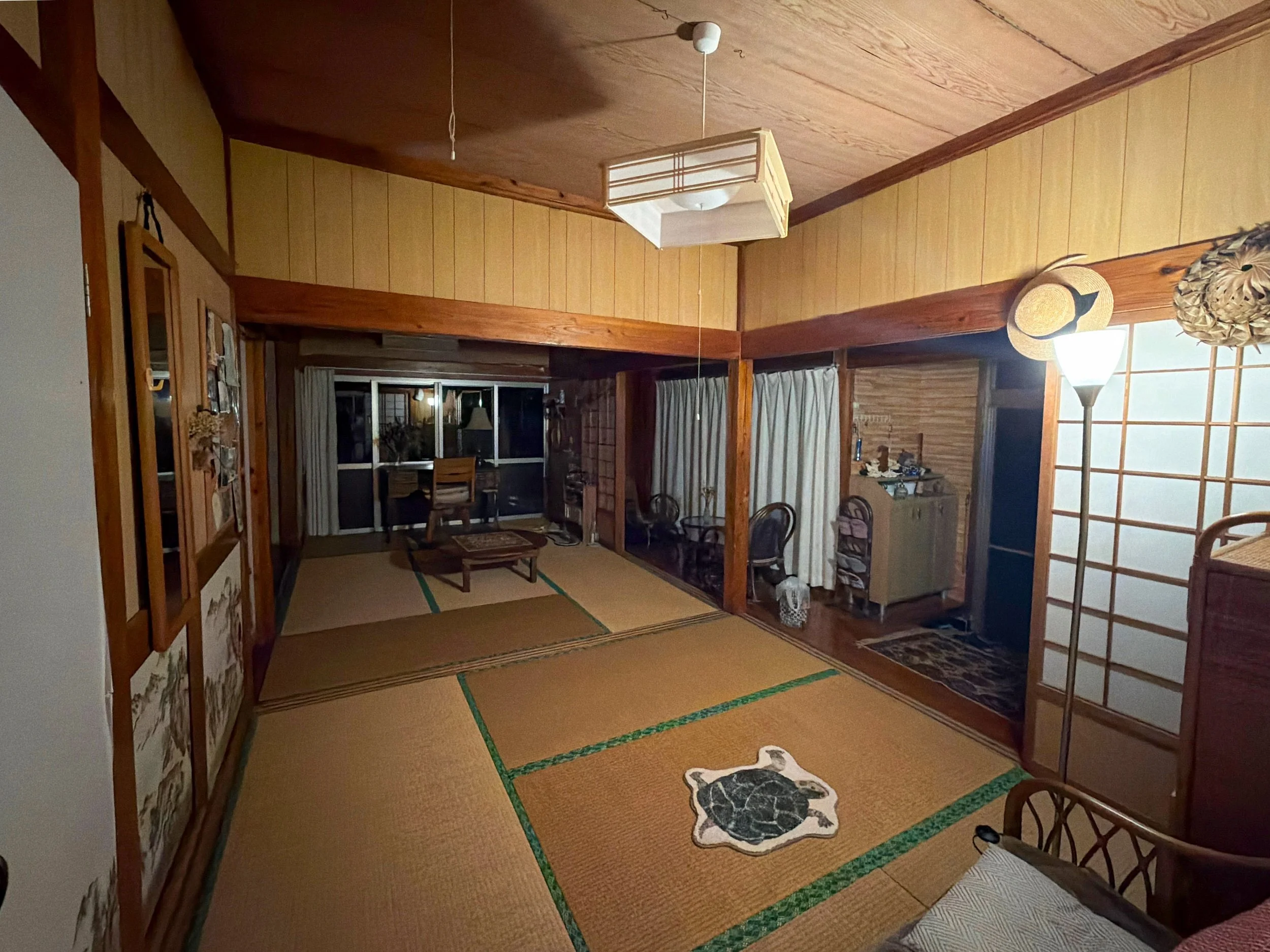 昔ながらの日本家屋の広い和室を写した室内です。床一面には縁が緑色の畳が敷かれ、中央には亀の絵が描かれた座布団のような敷物があります。太い木の柱や梁、木目の天井が温かみを感じさせます。奥には低い座卓と椅子、窓があり、右側には障子戸と食器棚があります。柔らかな照明が室内を照らし、落ち着いた懐かしい雰囲気が漂っています。