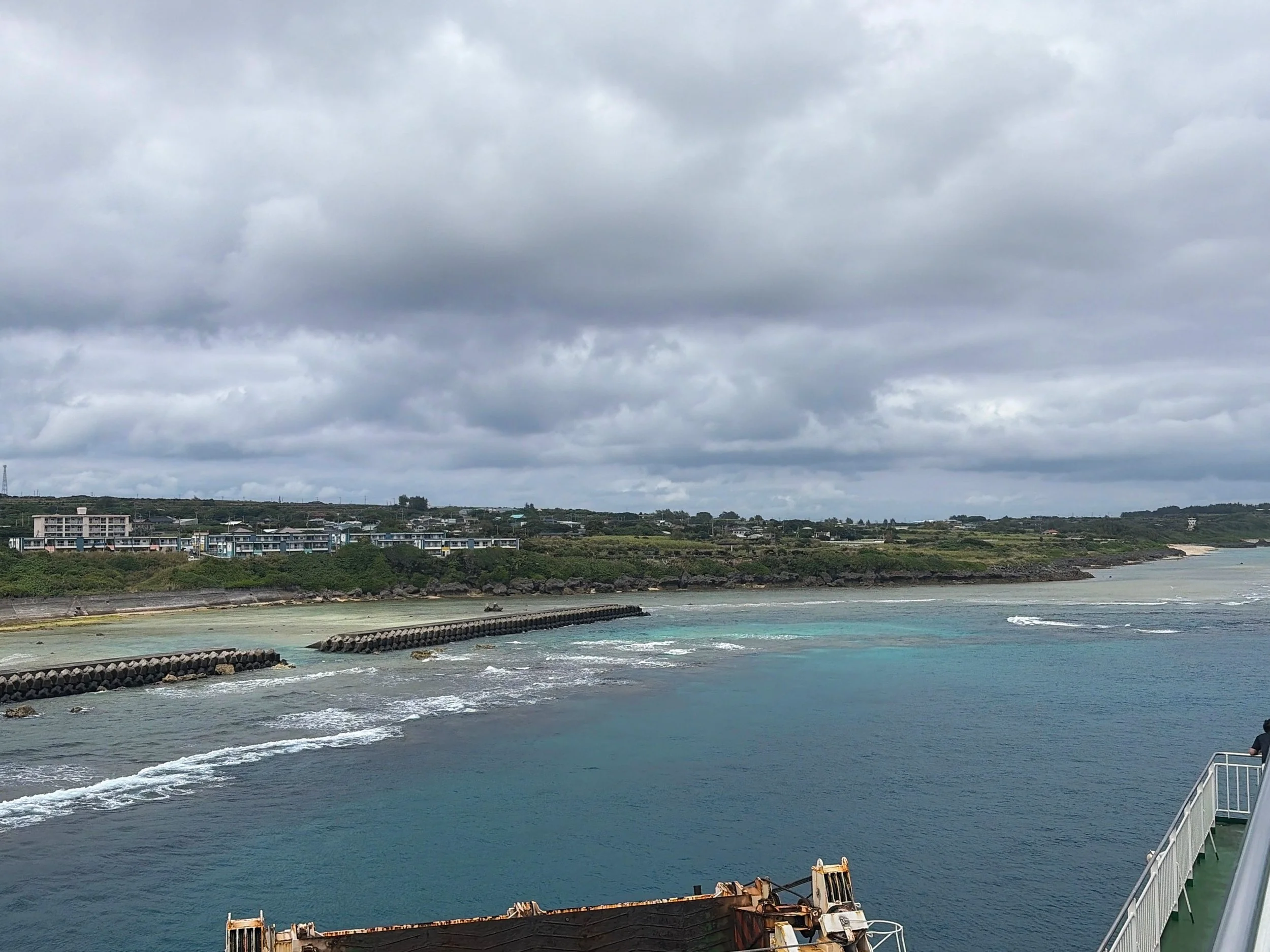 沖永良部島の和泊周辺を、船の上から眺めた風景です。手前には穏やかな青緑色の海が広がり、防波堤の周辺では白い波が細く打ち寄せています。対岸には緑豊かな丘と集落が広がり、住宅や建物が点在しています。遠くには砂浜も見えます。空一面には厚い灰色の雲が広がり、南の島らしい海と自然が静かに広がる景色です。
