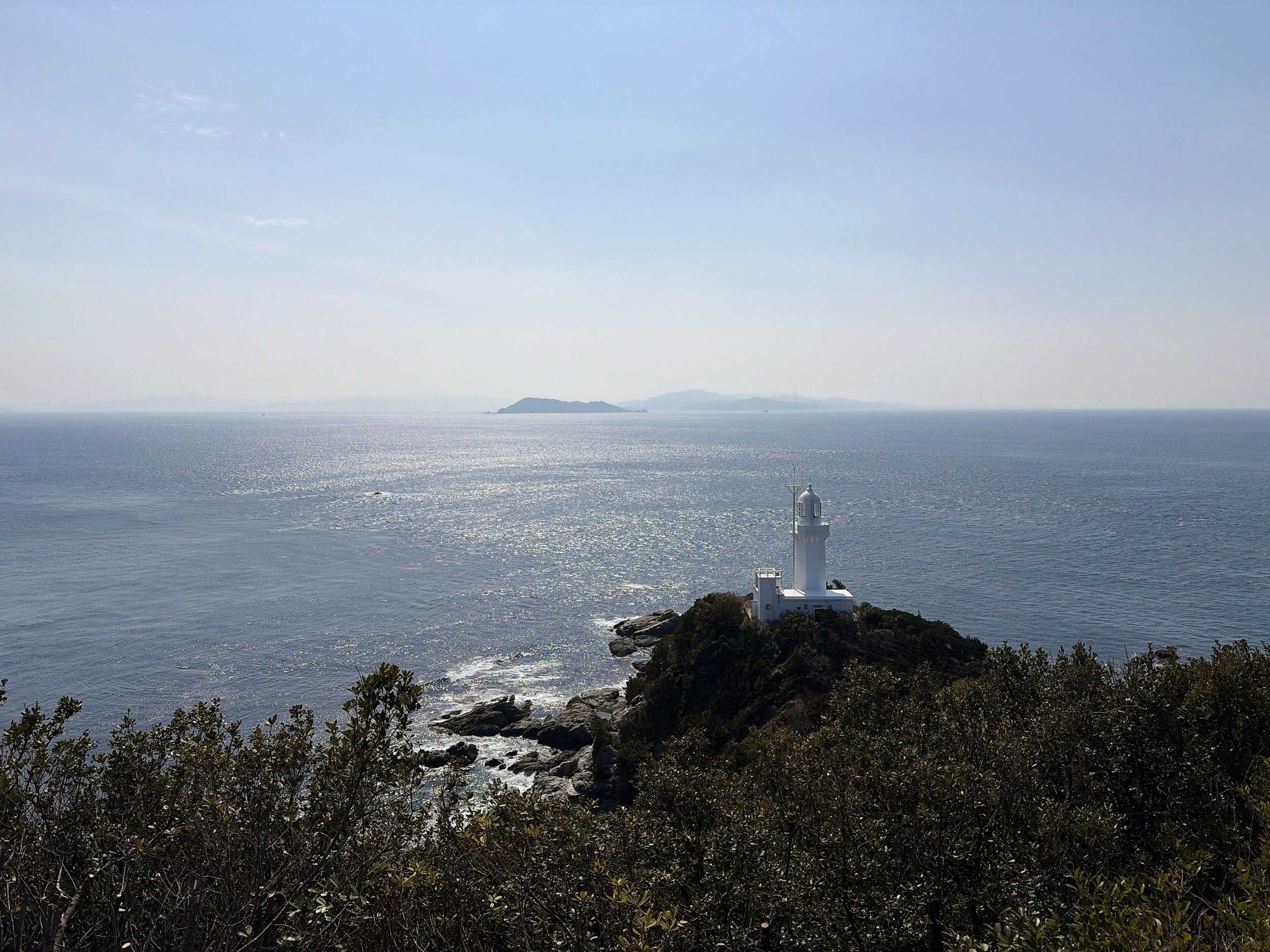 青空の下、海に突き出した岬の先端に白い灯台が建っています。灯台は緑の木々に覆われた小さな岬の上にあり、その周囲を岩場と穏やかな海が囲んでいます。海面には太陽の光が細かな銀色の輝きを作り、水平線の奥には霞んだ島々が浮かんでいます。手前には木々が広がり、青い海と白い灯台のコントラストが印象的な、開放感のある景色です。
