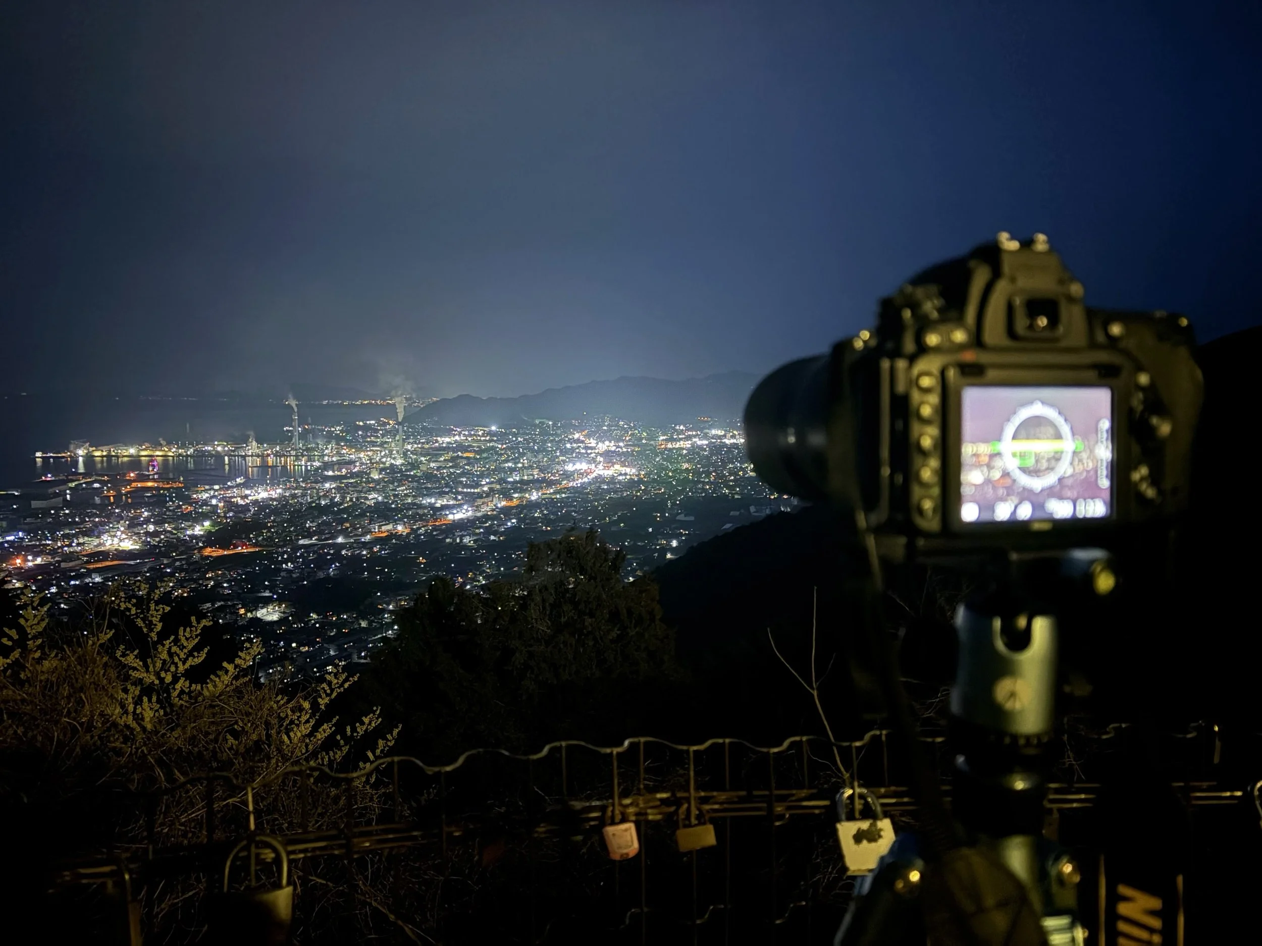 山の高台から見下ろした夜の街を撮影した写真です。遠くまで広がる街には無数の白やオレンジ色の明かりが輝き、左側には工場地帯と煙突、その奥には暗い山並みが見えます。手前右側には三脚に載せた一眼レフカメラがあり、カメラの向こうに広がる夜景を撮影しています。手前のフェンスには、色や形の異なる南京錠がいくつも掛けられています。