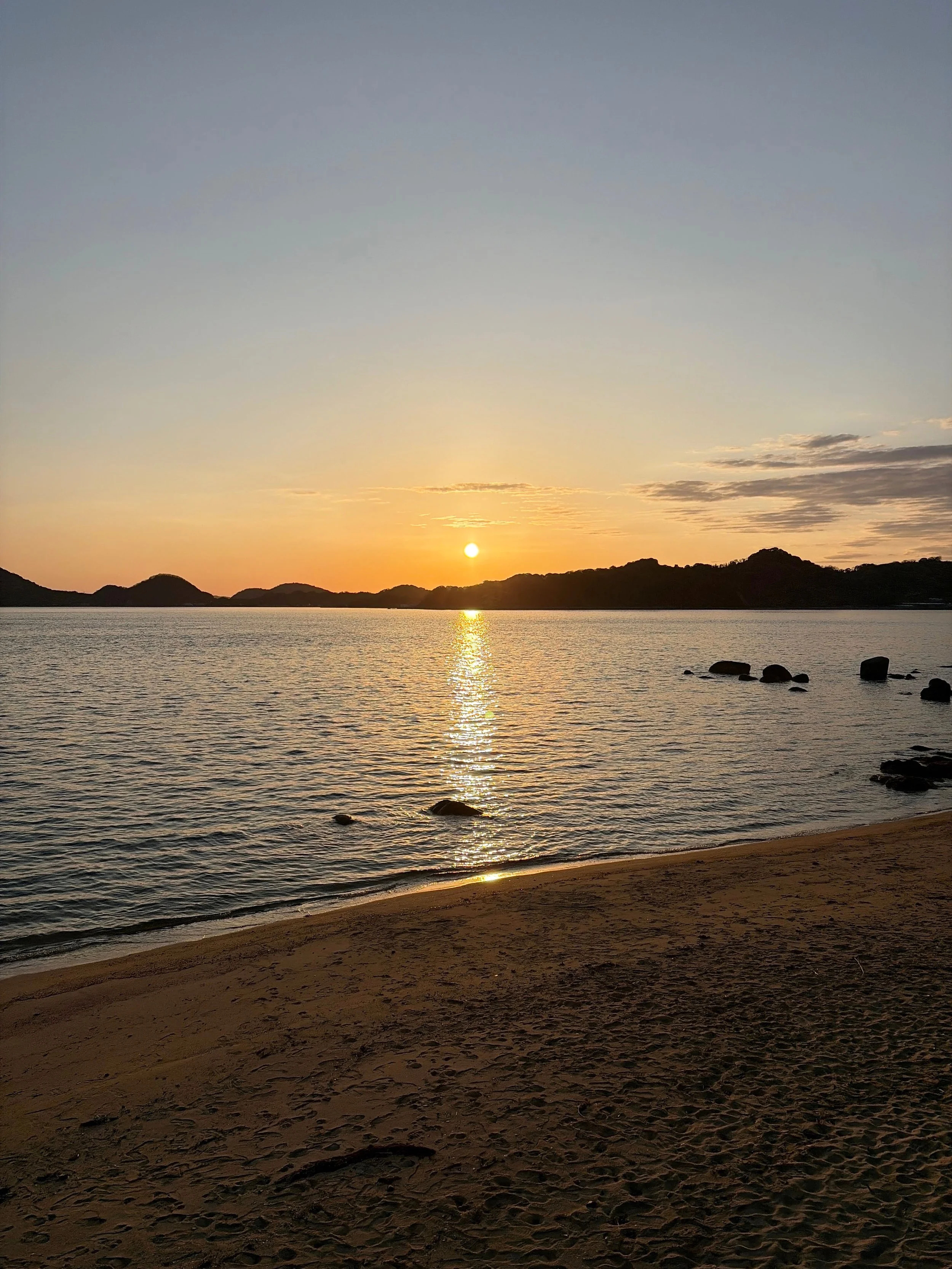 夕暮れの海辺を写した風景です。水平線近くにオレンジ色の太陽が浮かび、海面には太陽から手前へ細長く輝く光の道が伸びています。遠くには丸みを帯びた島や山々が黒いシルエットとなって連なり、右側の海にはいくつかの岩が浮かんでいます。手前には砂浜が広がり、穏やかな波が静かに打ち寄せています。空は淡い橙色から灰青色へ変化し、静かな夕暮れを感じさせます。