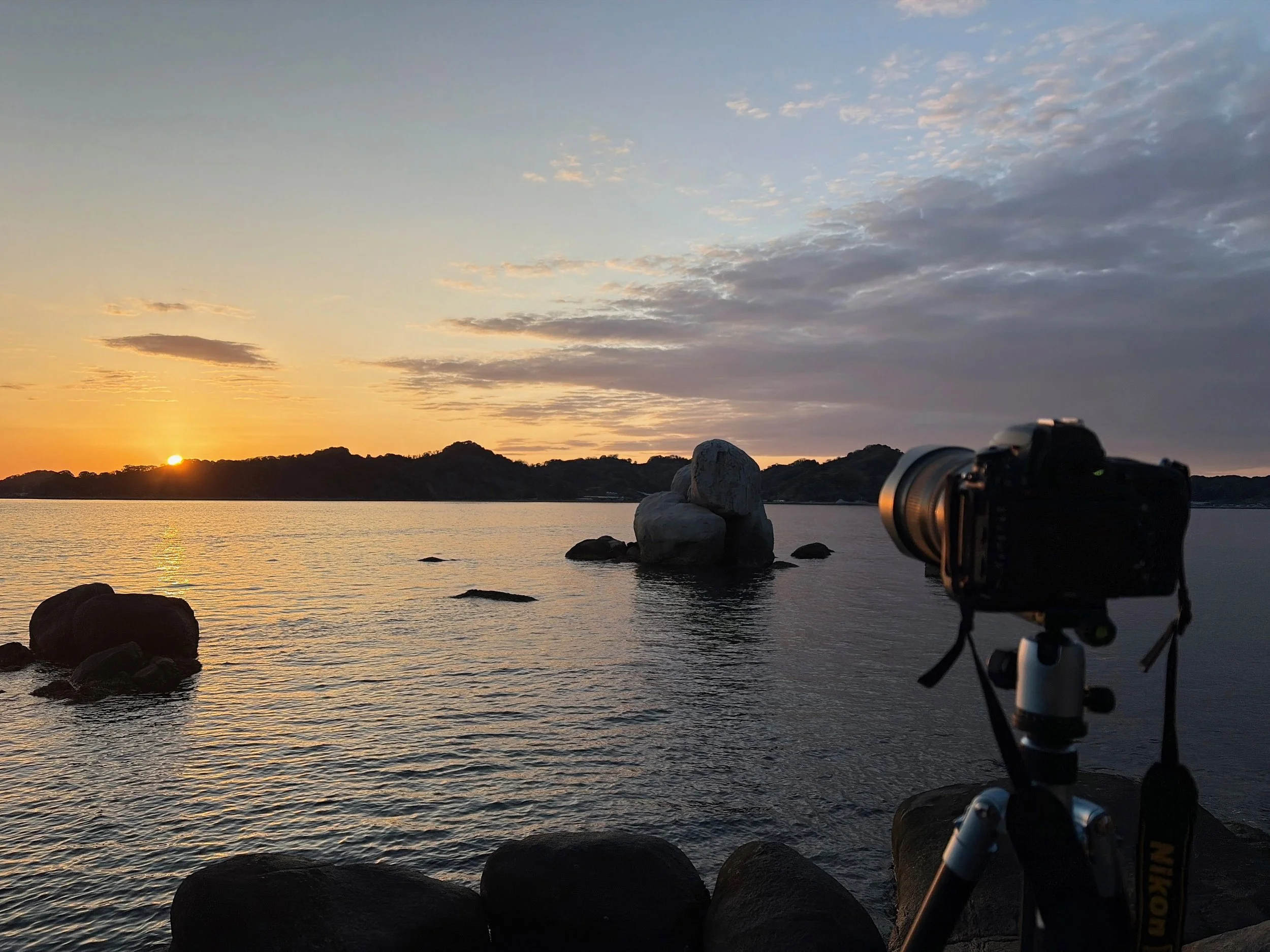 夕暮れの海辺を撮影した一枚です。水平線の左側に沈みかけた太陽があり、海面には黄金色の光が伸びています。中央には大きな丸い岩が重なるように海から突き出し、周囲にも大小の岩が点在しています。右手前には三脚に固定された一眼レフカメラが海へ向けられています。空には薄い雲が広がり、夕焼けの橙色と青灰色が混ざった幻想的な景色です。