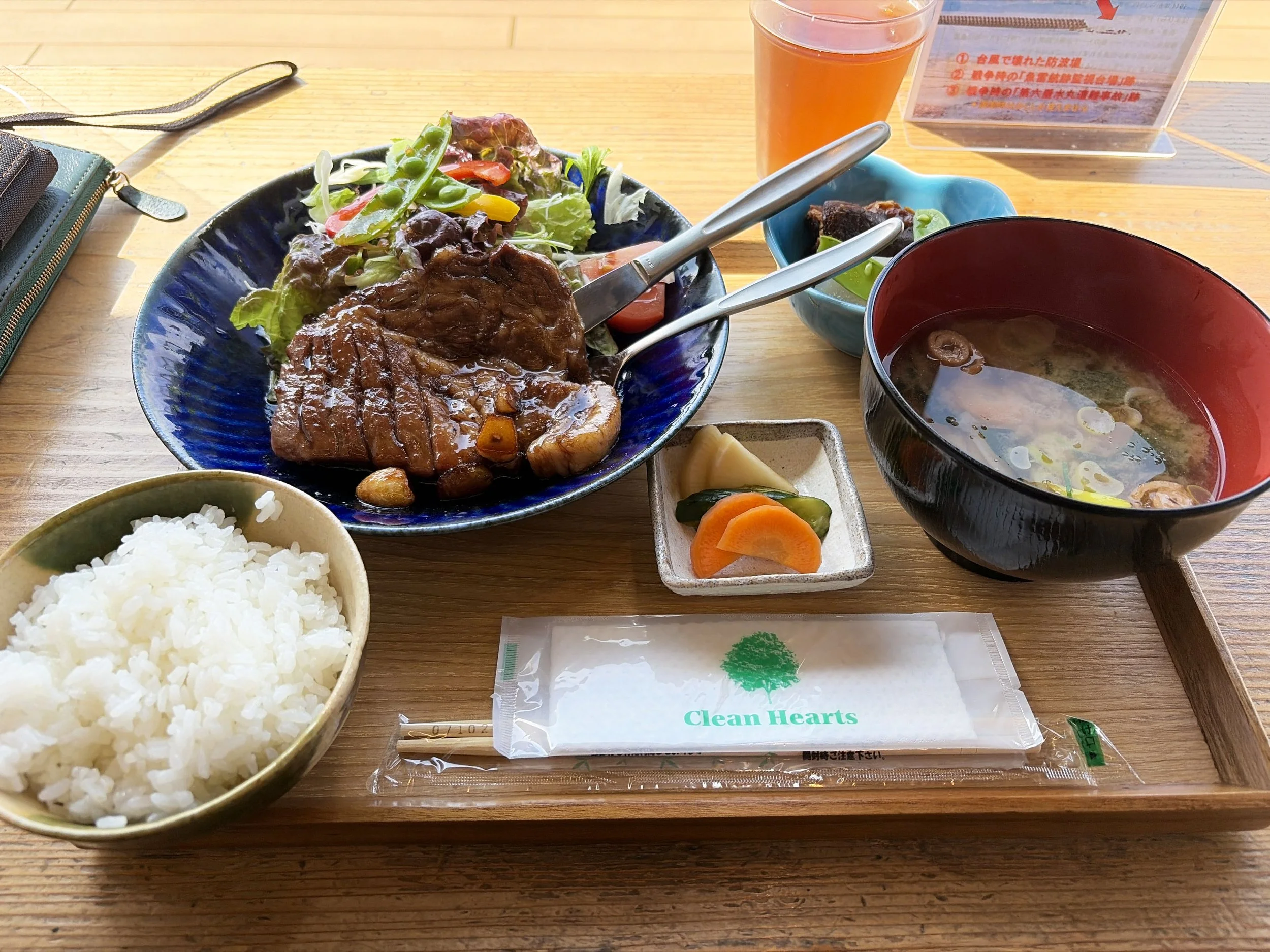 木目のテーブルの上に、和食の定食が並んでいます。中央には照りのある厚切りのステーキが盛られ、奥には色鮮やかな野菜サラダがあります。左手前には白いご飯、右側には赤と黒の器に入った味噌汁。中央手前には、きゅうりやにんじんなどの漬物も添えられています。奥にはオレンジ色の飲み物があり、箸やスプーンも置かれています。全体的に温かみのある、ボリュームたっぷりの食事です。